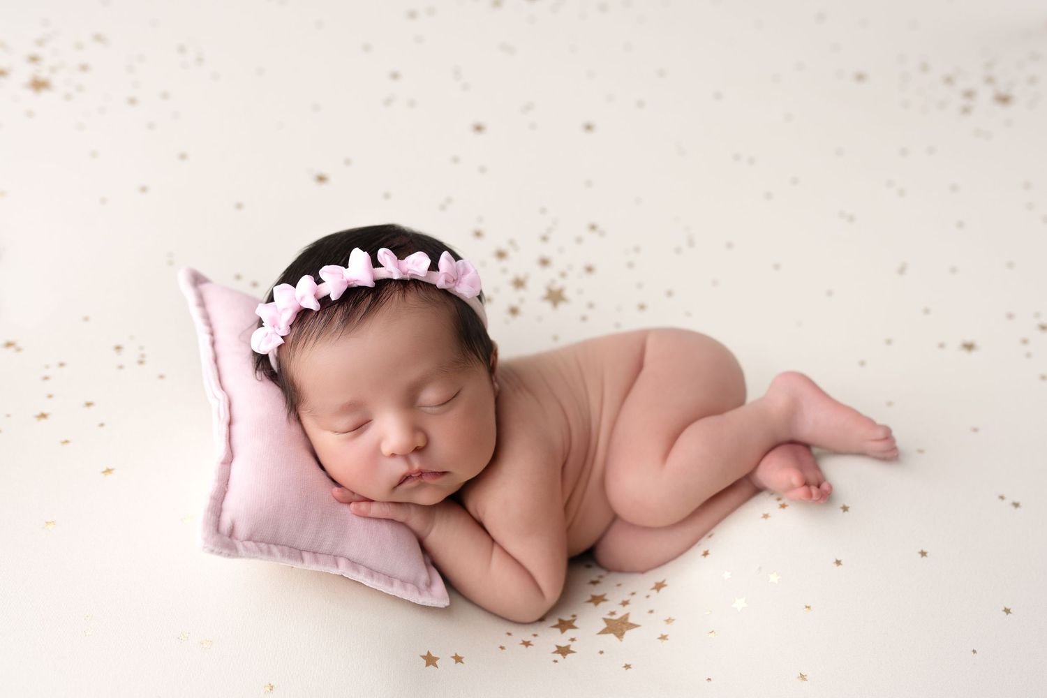Newborn baby sleeping peacefully on white blanket wearing delicate pink crown headpiece.