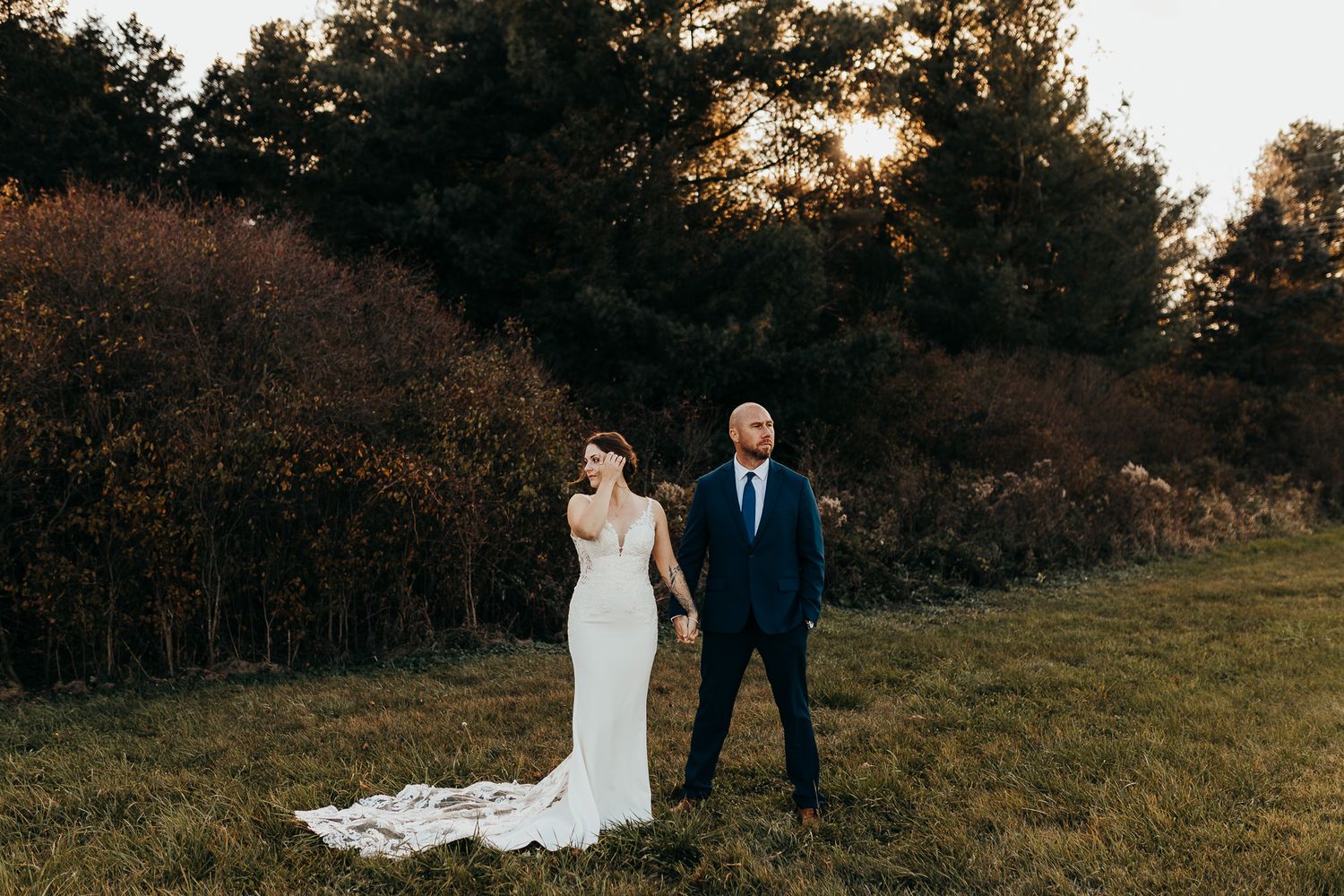 A couple walks through a sunset-lit meadow with autumn trees in the background during their wedding photoshoot.