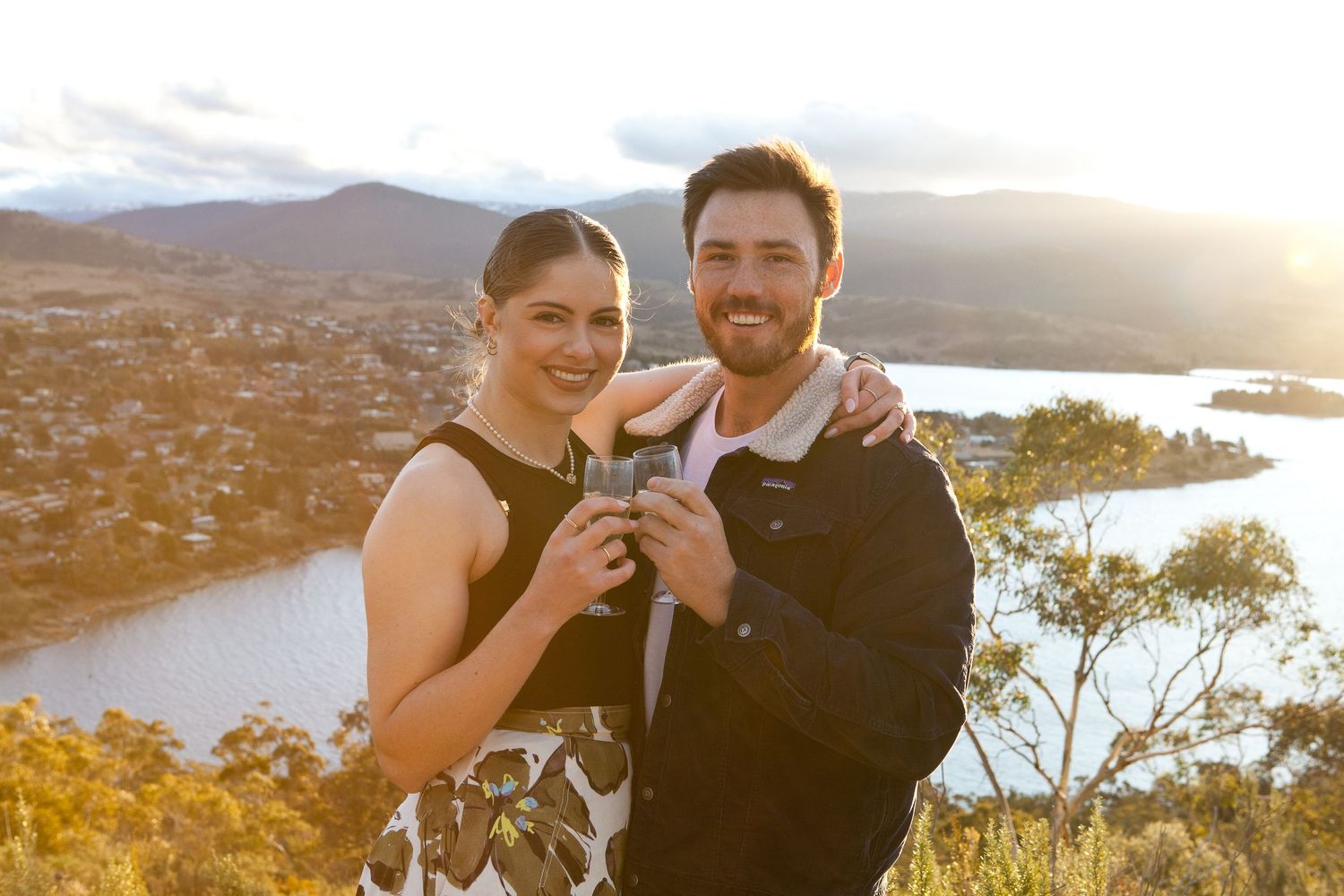 Couple embraces on hilltop overlooking scenic bay at golden sunset after proposal.