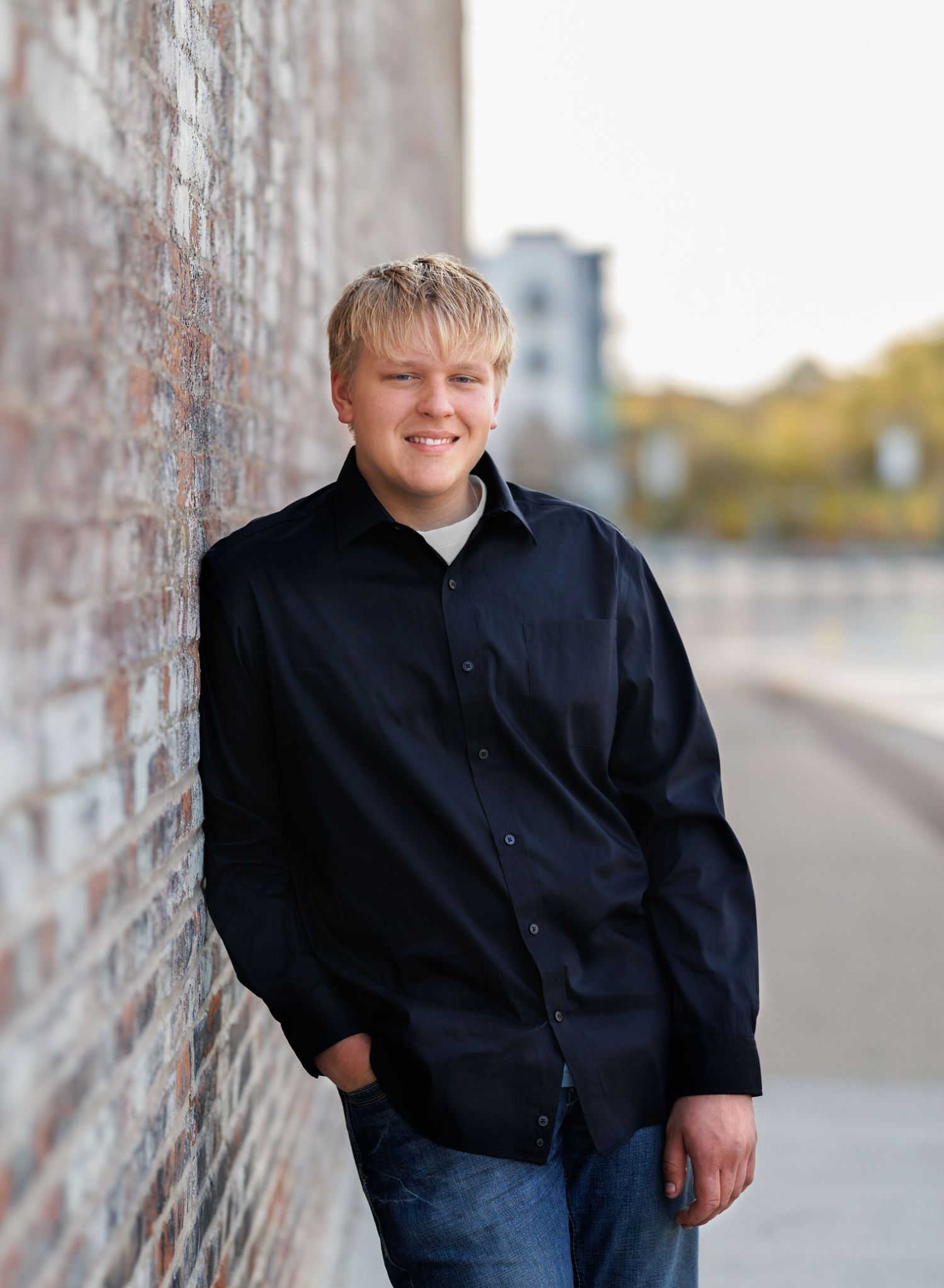 A senior boy in a black button-down shirt leans against a brick wall outdoors on an autumn day.