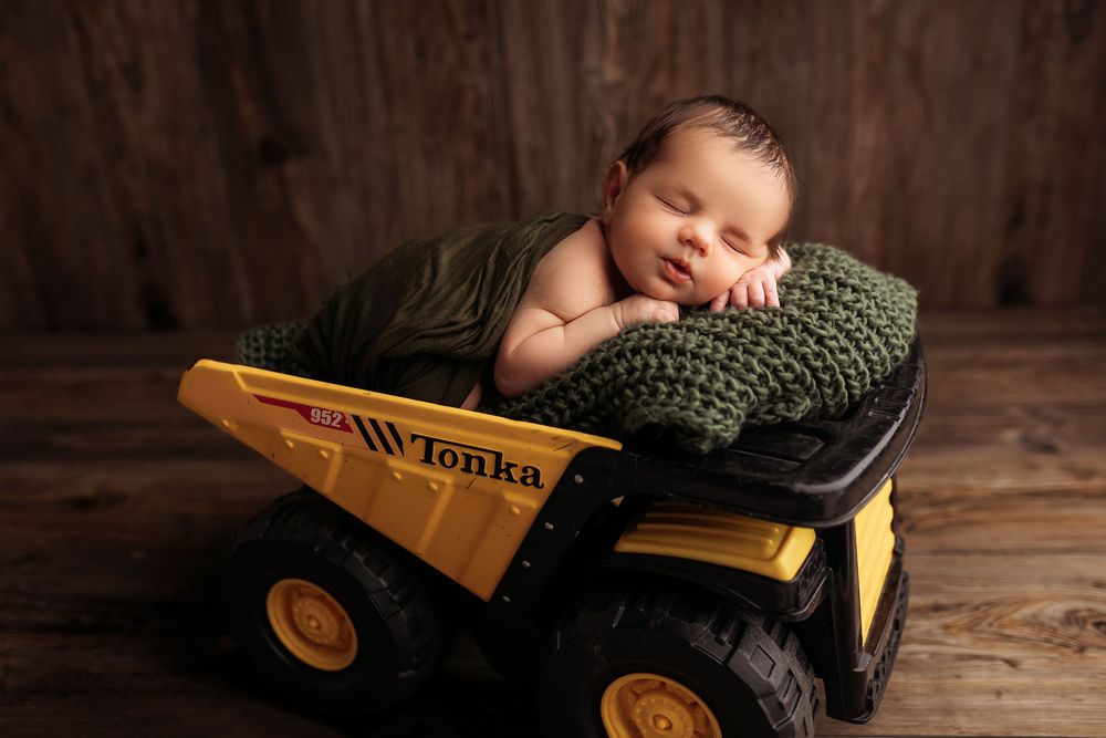A newborn baby sleeps peacefully while nestled against a toy construction truck in a rustic wooden setting.