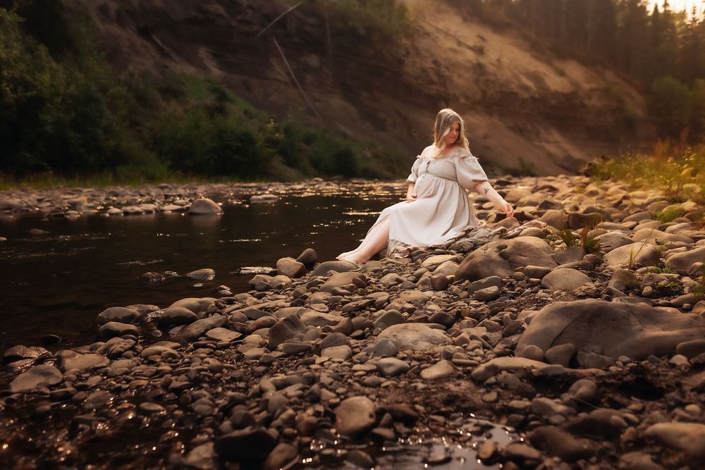 A person in a flowing white dress sits gracefully on rocky terrain by a river at sunset.