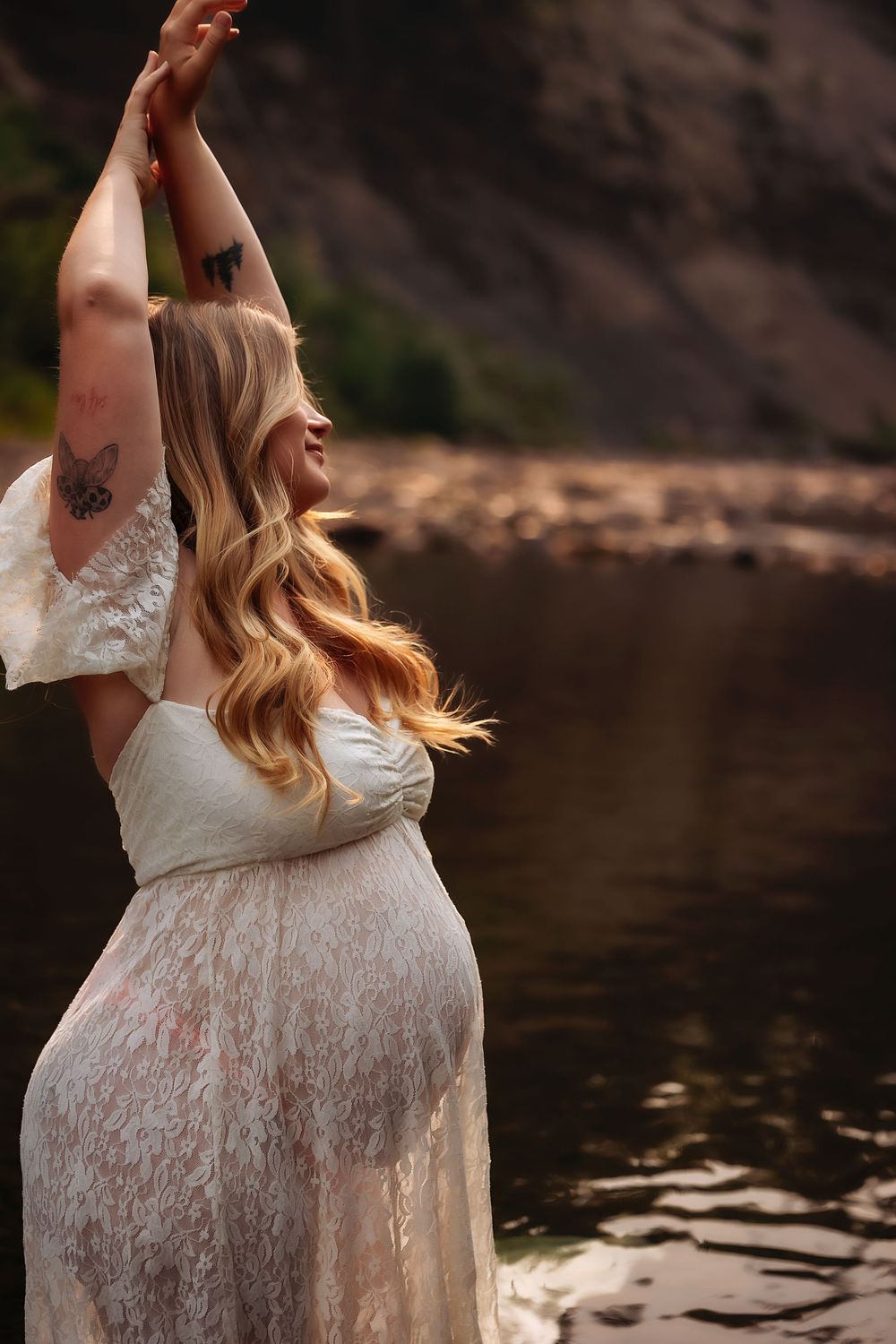 Woman in white lace dress poses gracefully by a serene lake during a golden hour photoshoot.