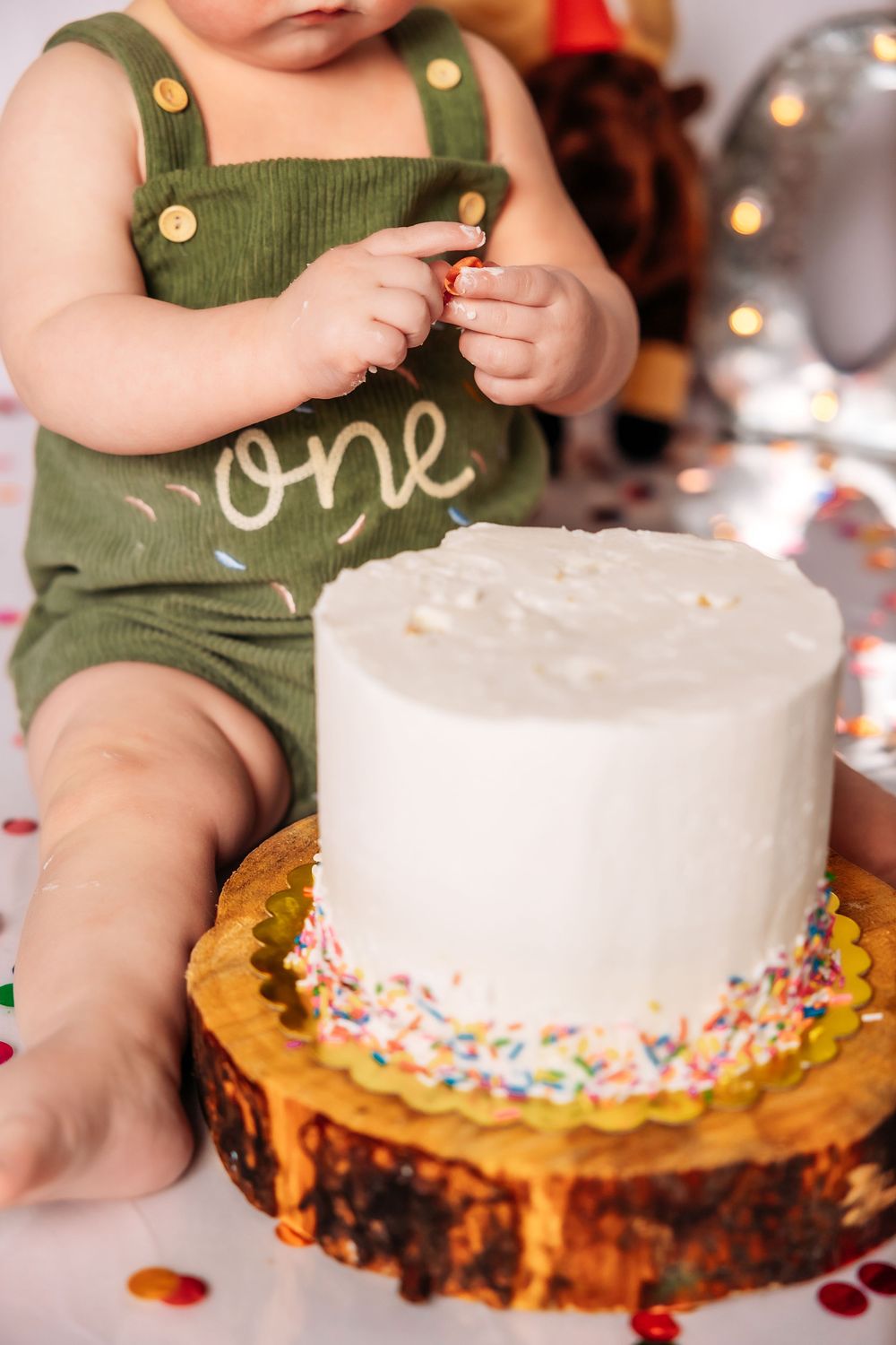 A white frosted cake with rainbow sprinkles sits on a wooden surface as small hands reach to touch it.