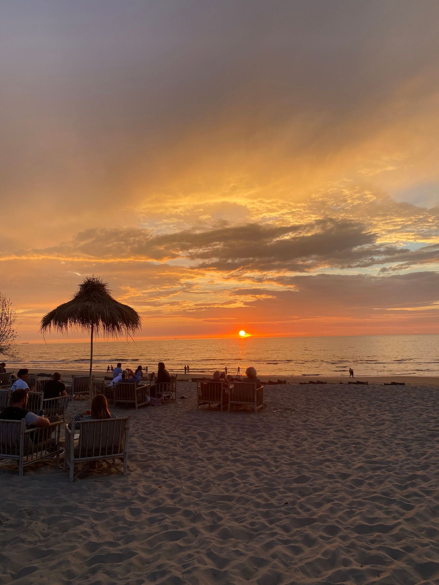 People relaxing on sandy beach under thatched umbrella watching a vibrant orange and red sunset over the ocean.