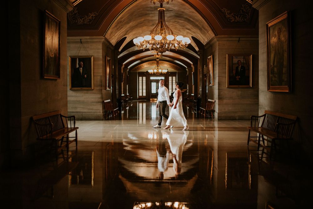 Engaged couple walks through a historical building holding hands with details such as their reflection in the photo.