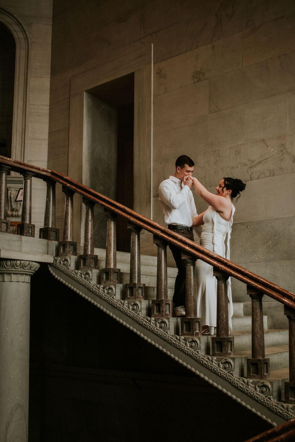 Fiancé kisses his soon to be wife's hand as they stand on the staircase leading up while their affordable photographer takes a photo.