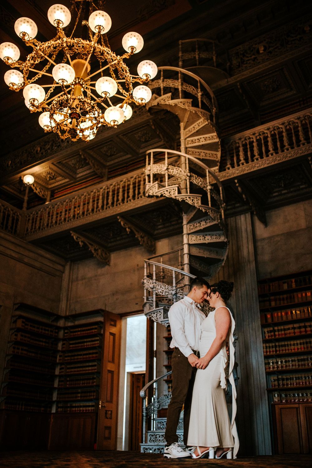 Engaged couple in a historical room in the state capital with a winding staircase and a ton of books.