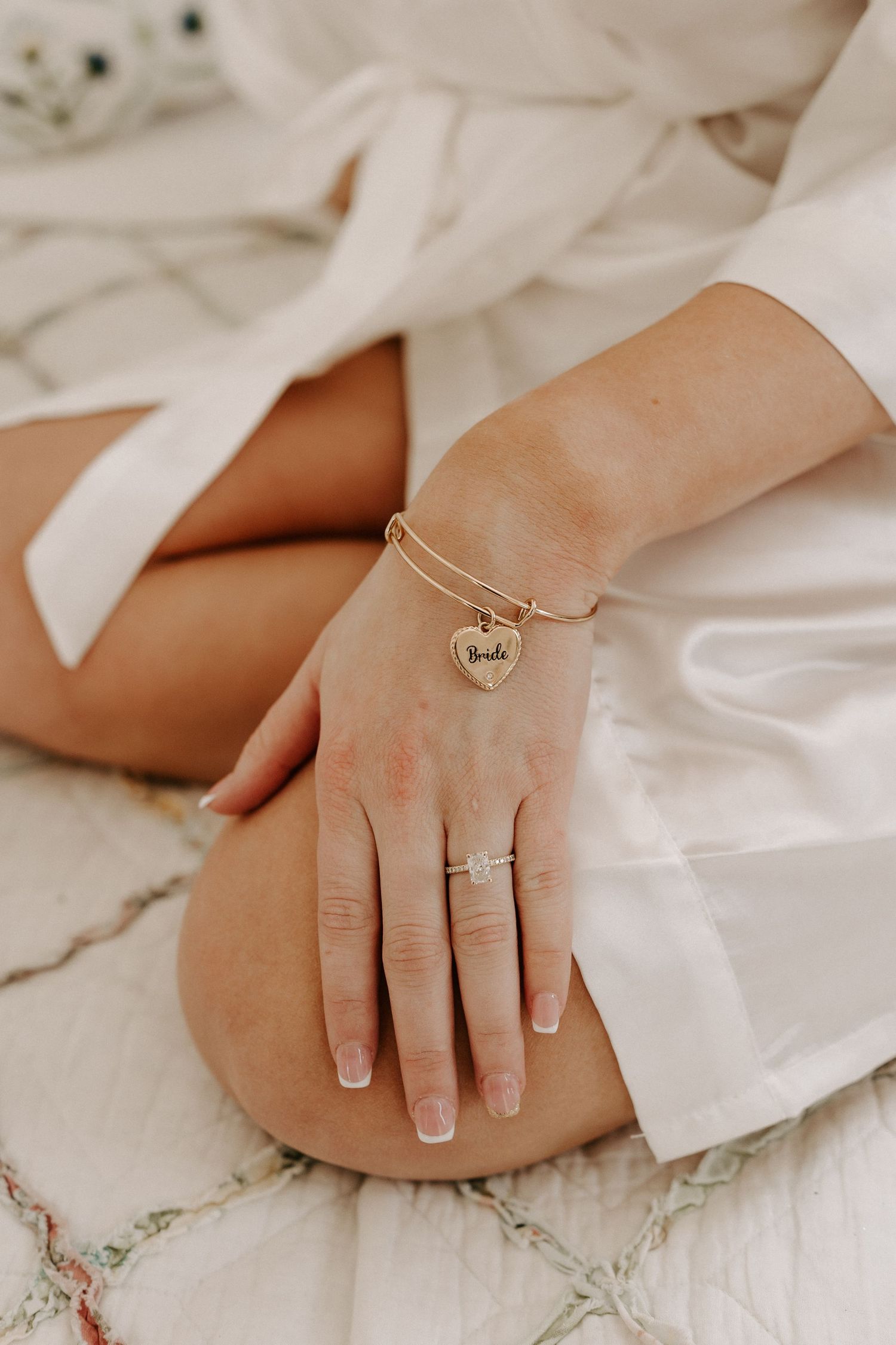 Bride wearing gold heart charm bracelet and engagement ring in white bridal robe.