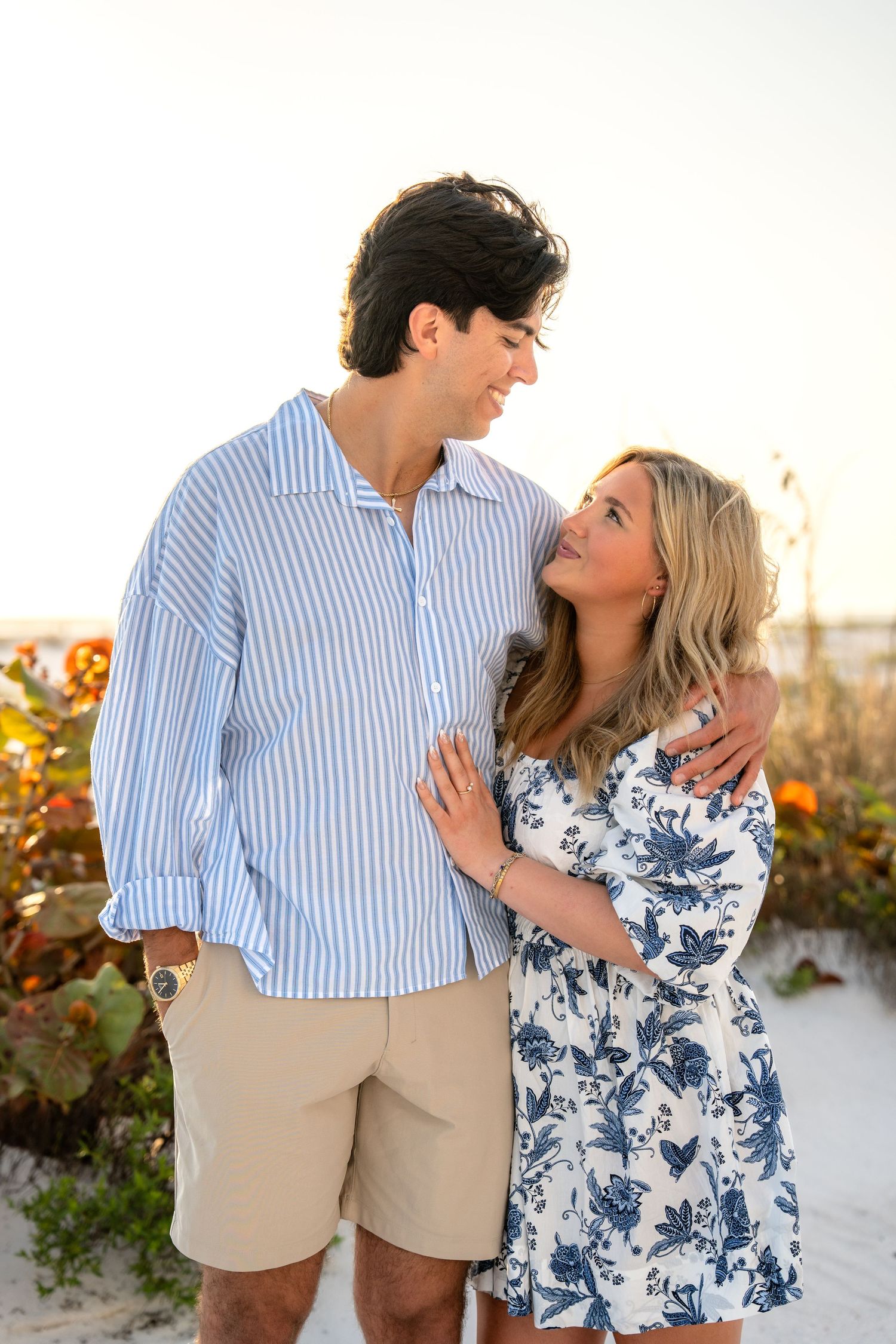 A young couple gazes at each other lovingly on a white sand beach at golden hour among coastal vegetation.