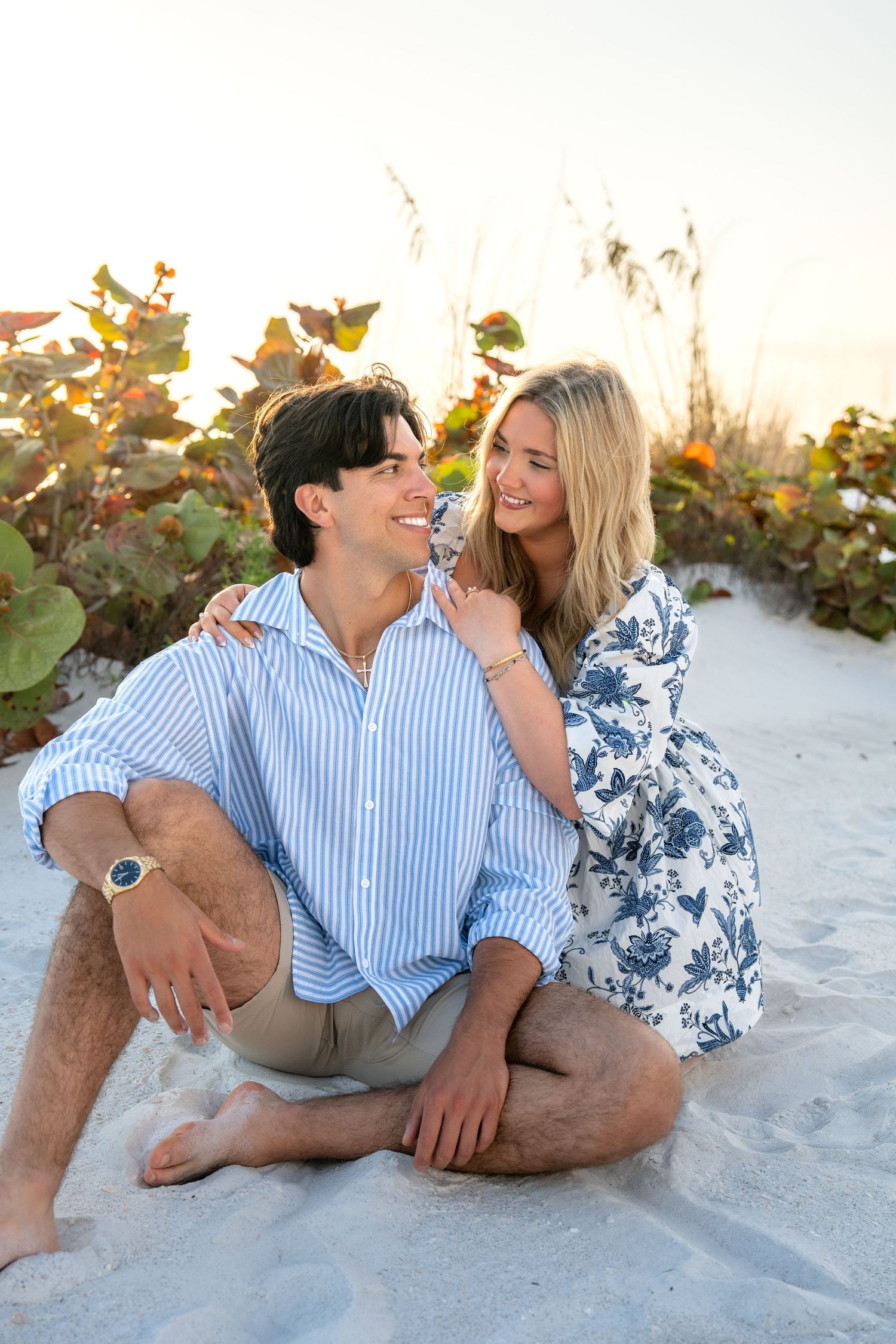 A couple sits intimately on white sand at golden hour, smiling and embracing among colorful beach vegetation.