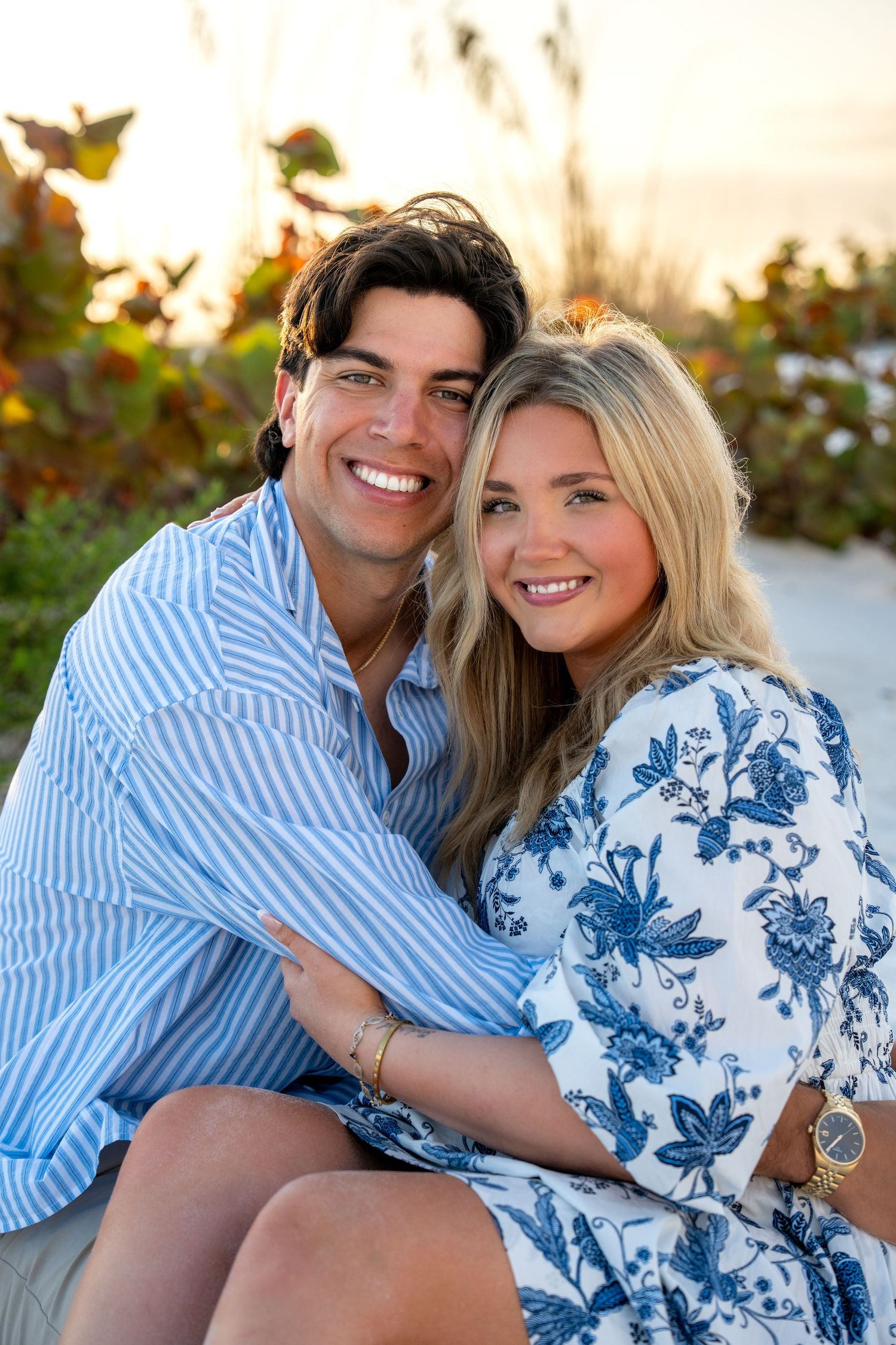 Smiling couple posing together on a beach at sunset, wearing coordinating blue and white outfits.