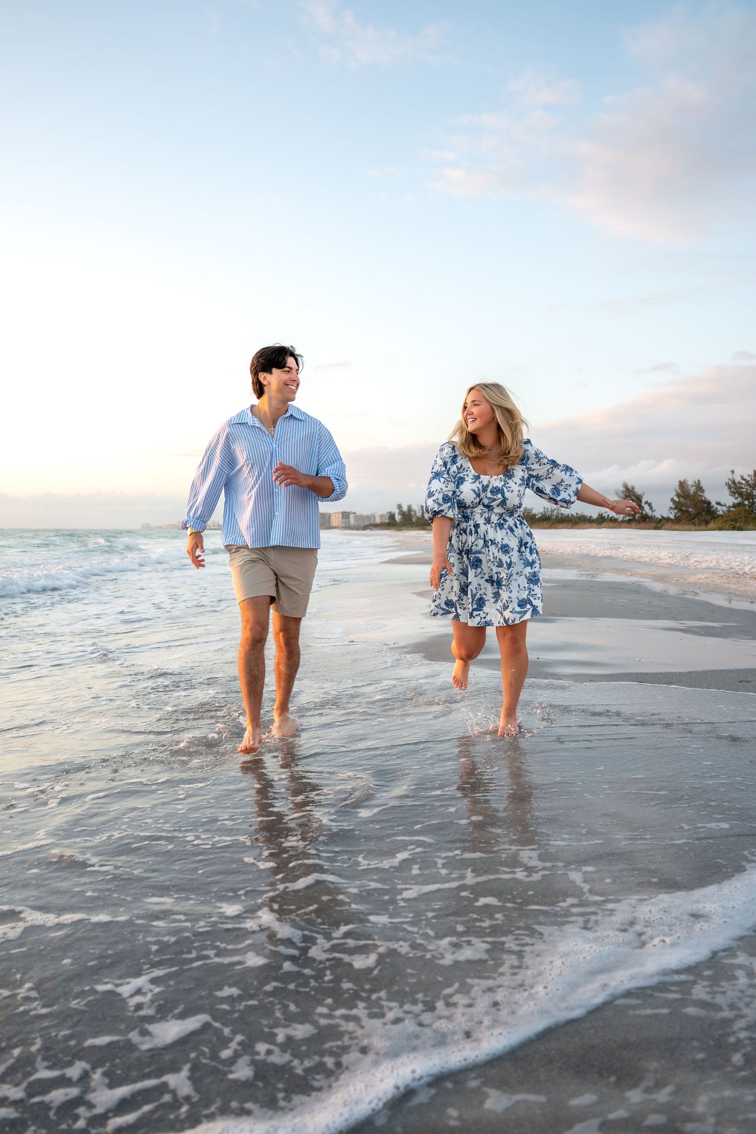 Happy couple walking barefoot on a beach at sunset, splashing through shallow ocean waves together.