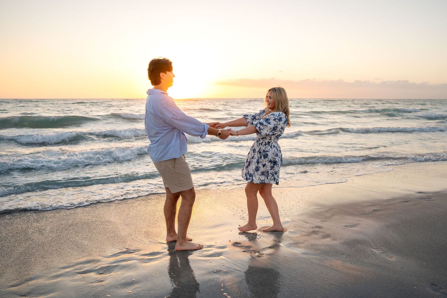 Couple holding hands facing each other on a beach at golden sunset with ocean waves behind them.