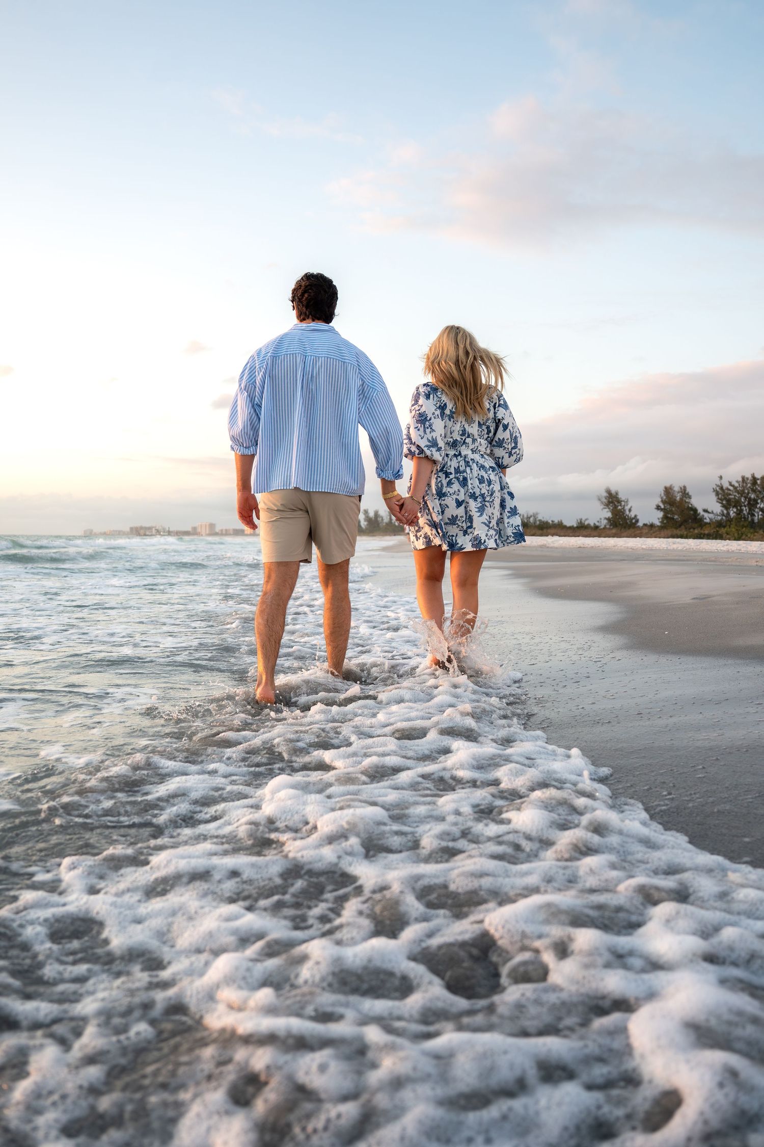 Couple holding hands walking away along a beach shoreline with foamy waves and pastel sunset sky.