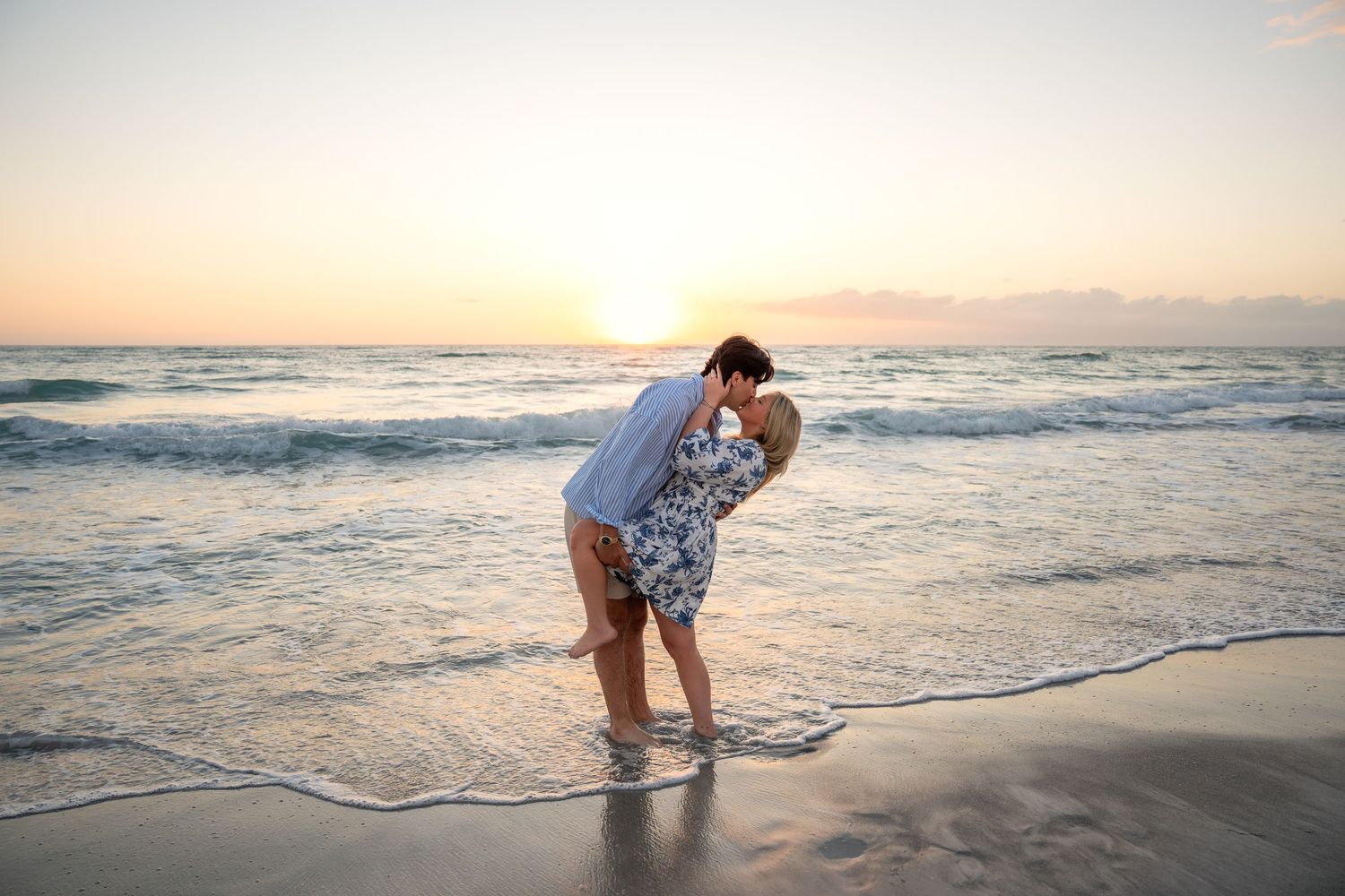 Couple sharing a romantic dip kiss on a sandy beach at sunset with golden ocean backdrop.