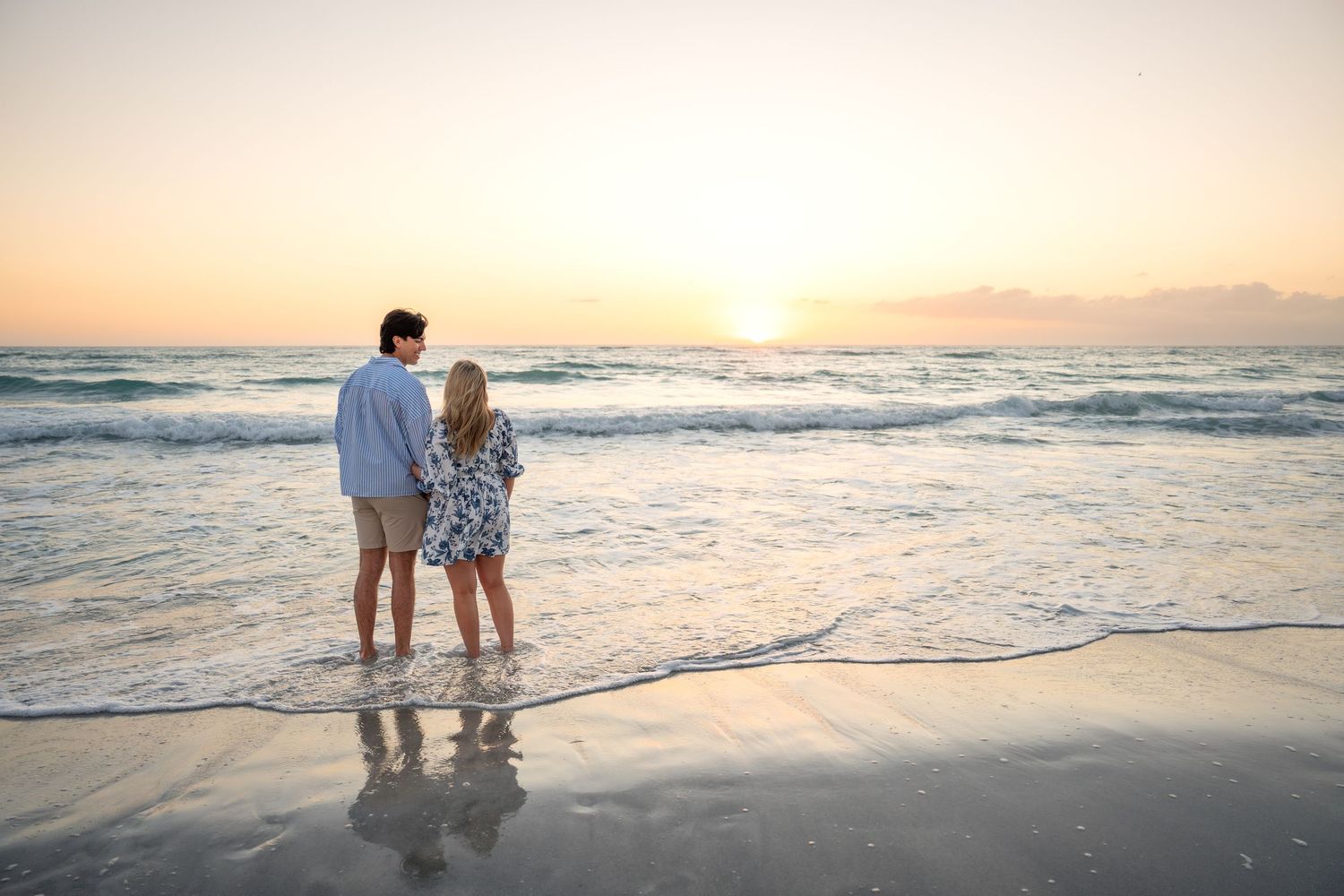 Couple stands hand-in-hand at shoreline watching a golden sunset over calm ocean waves.