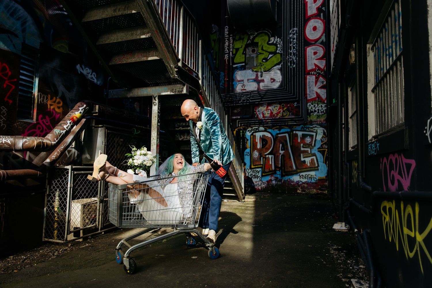 Creative Auckland wedding photo of blue-haired bride & groom in velvet jacket in a laneway with lots of graffiti.