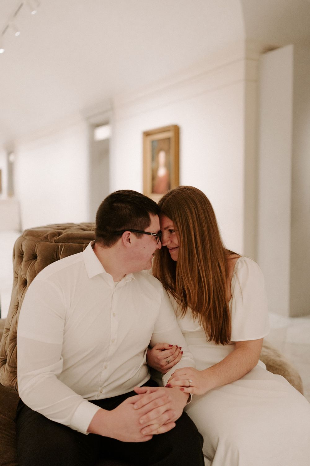 Couple sitting close together on tufted velvet bench in art gallery, smiling and holding hands.