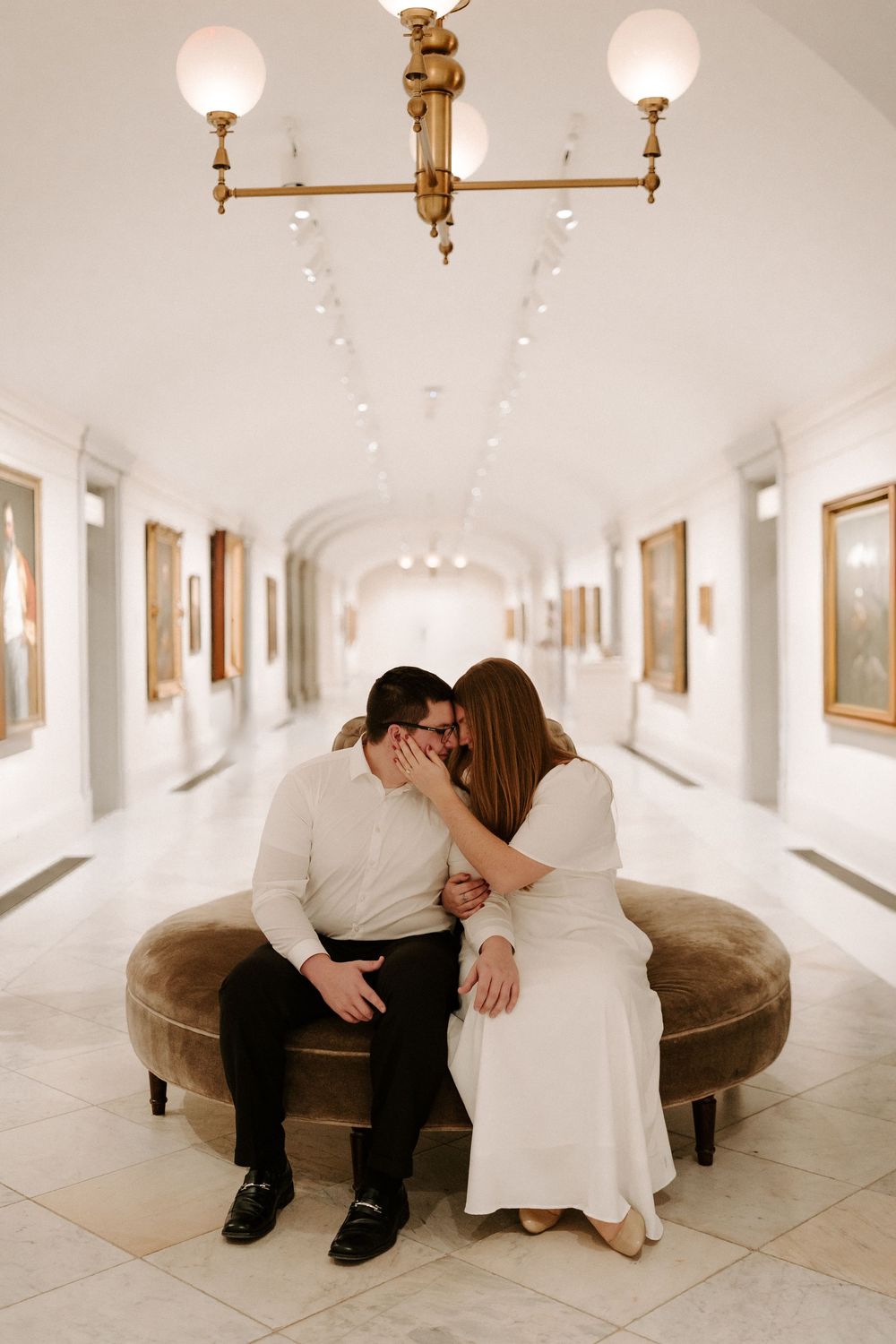 Couple sharing a kiss on round velvet bench in elegant museum hallway with gold chandelier.