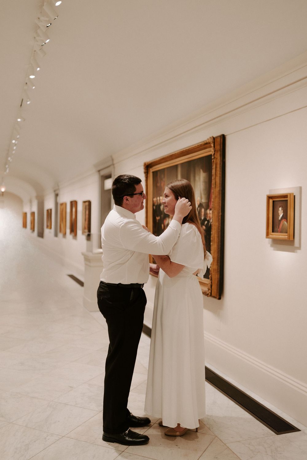 Man tenderly brushing woman's hair back as they stand together in museum gallery hallway.