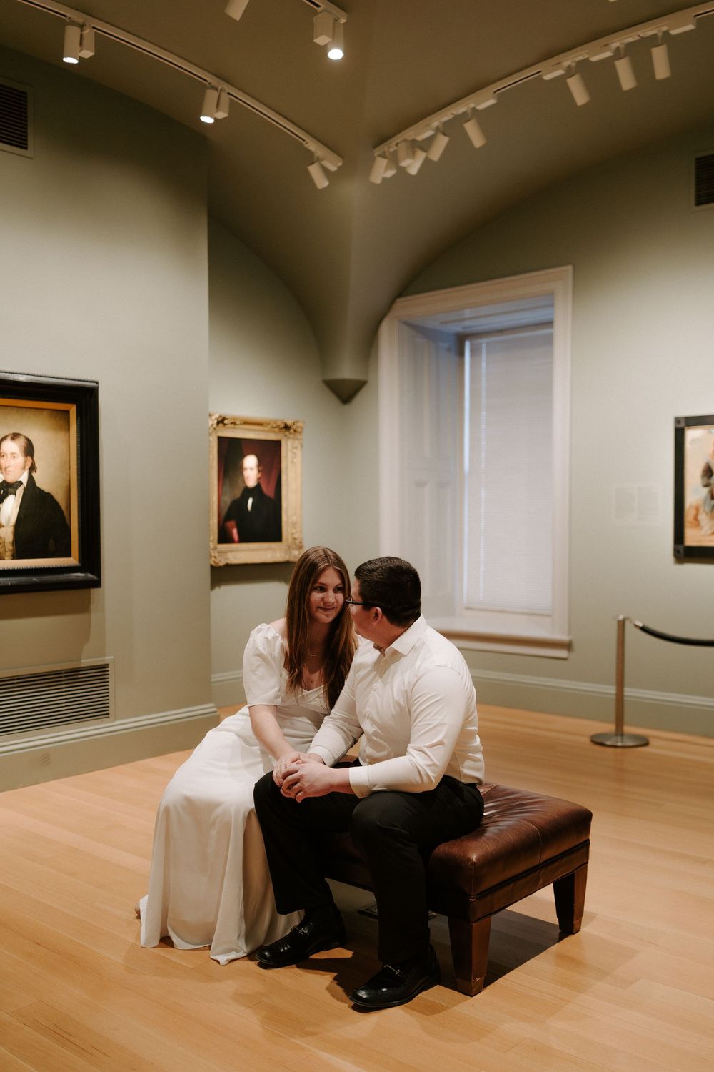 Couple sitting intimately on leather bench in art museum gallery surrounded by portrait paintings.