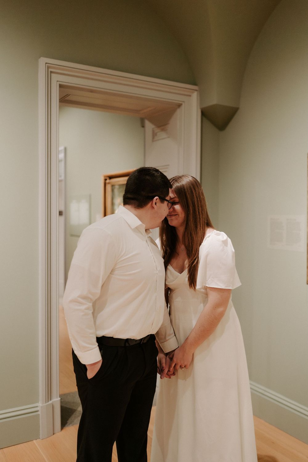 Couple holding hands and sharing a forehead kiss in soft green museum gallery doorway.