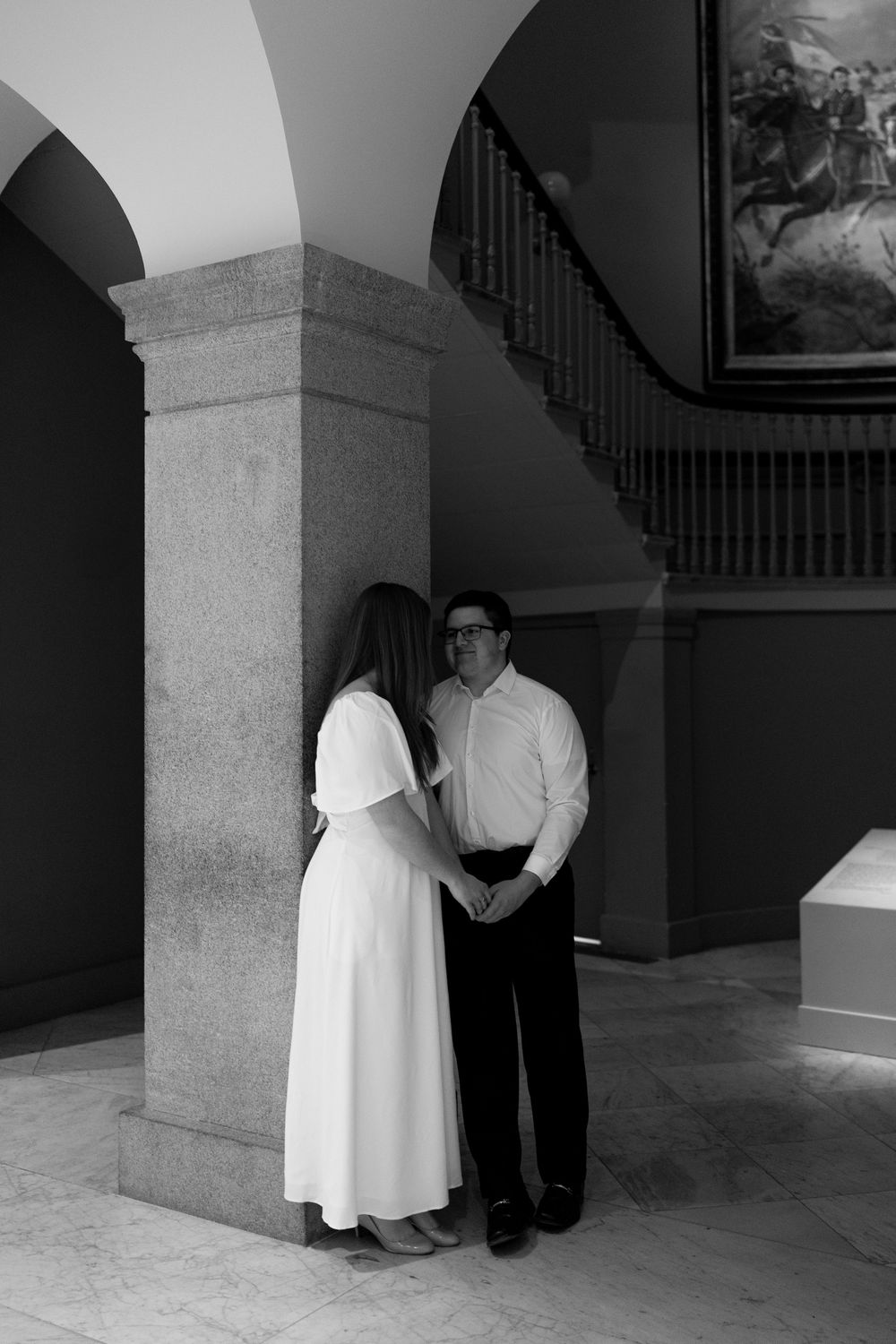 Black and white photo of couple holding hands behind stone column with grand staircase backdrop.