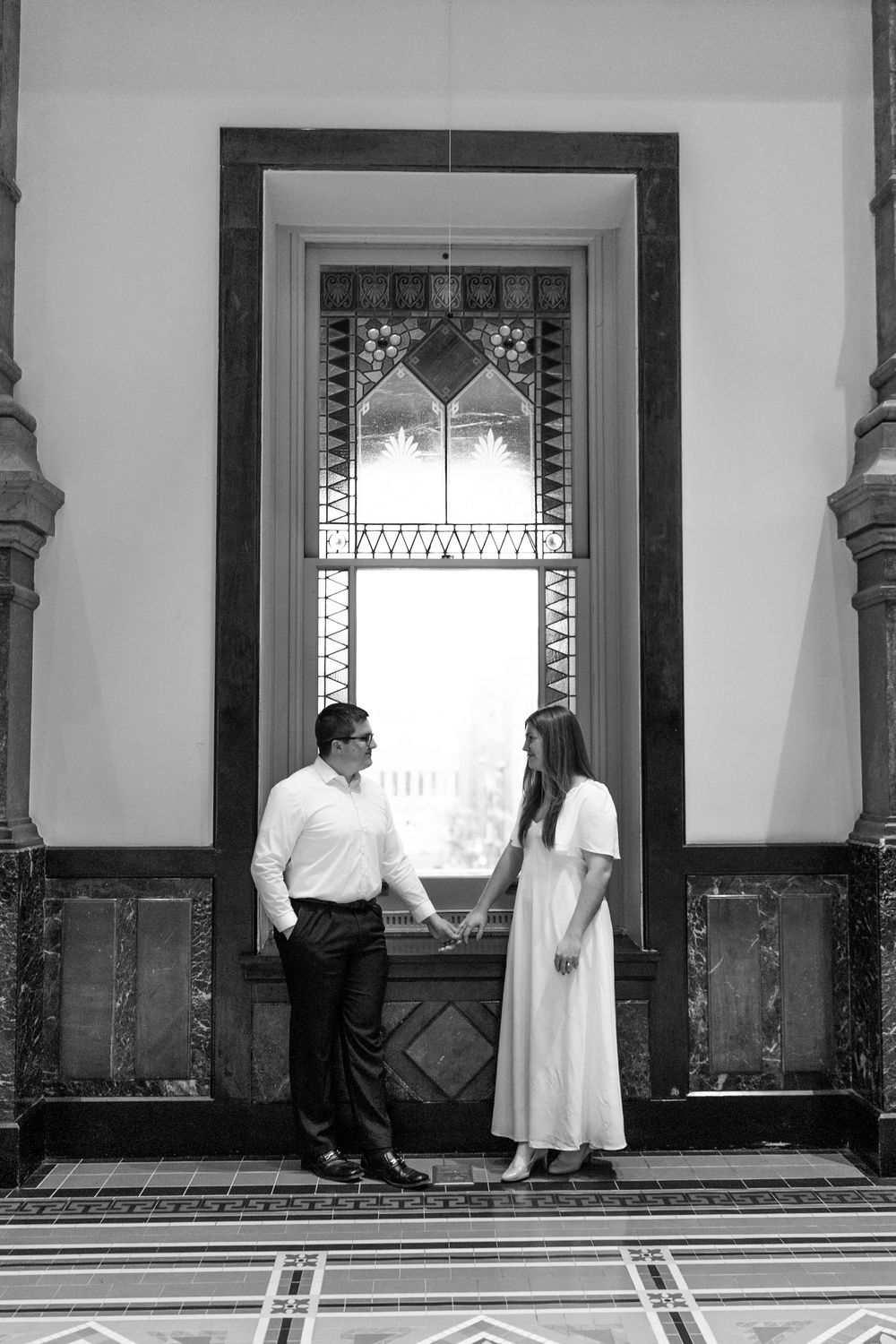 Black and white photo of couple holding hands before ornate stained glass window in historic building.