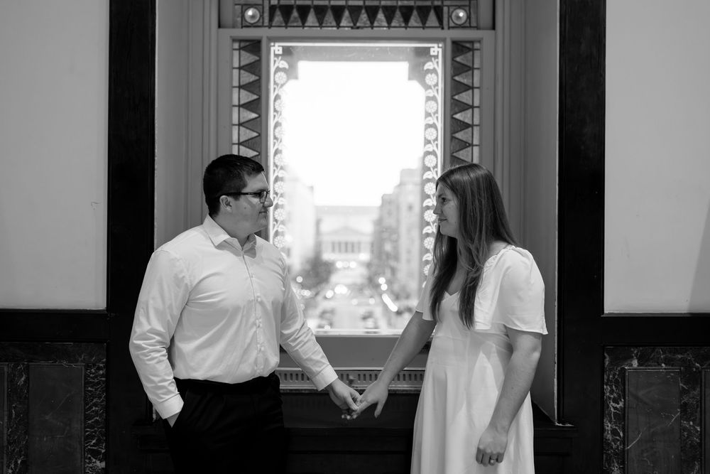 Black and white photo of couple holding hands by ornate window with city view in historic building.