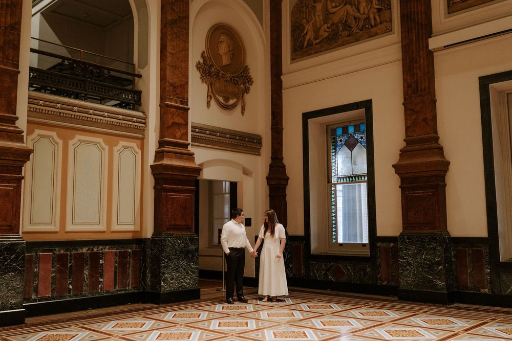 Couple holding hands in grand ornate hall with decorative mosaic tile floors and marble columns.