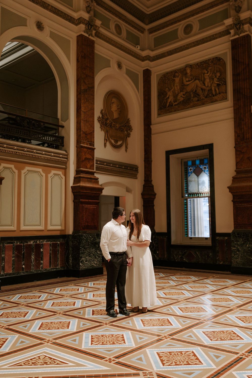 Couple standing together in stunning historic building with intricate tile floors and classical murals.