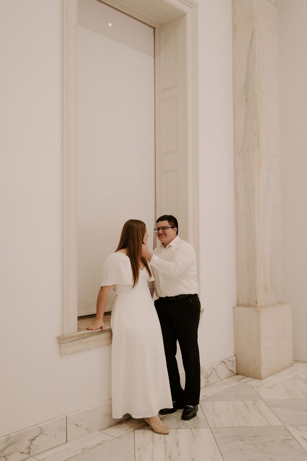 Couple sharing a tender moment in a marble hallway, woman in white dress and man in white shirt and black pants.