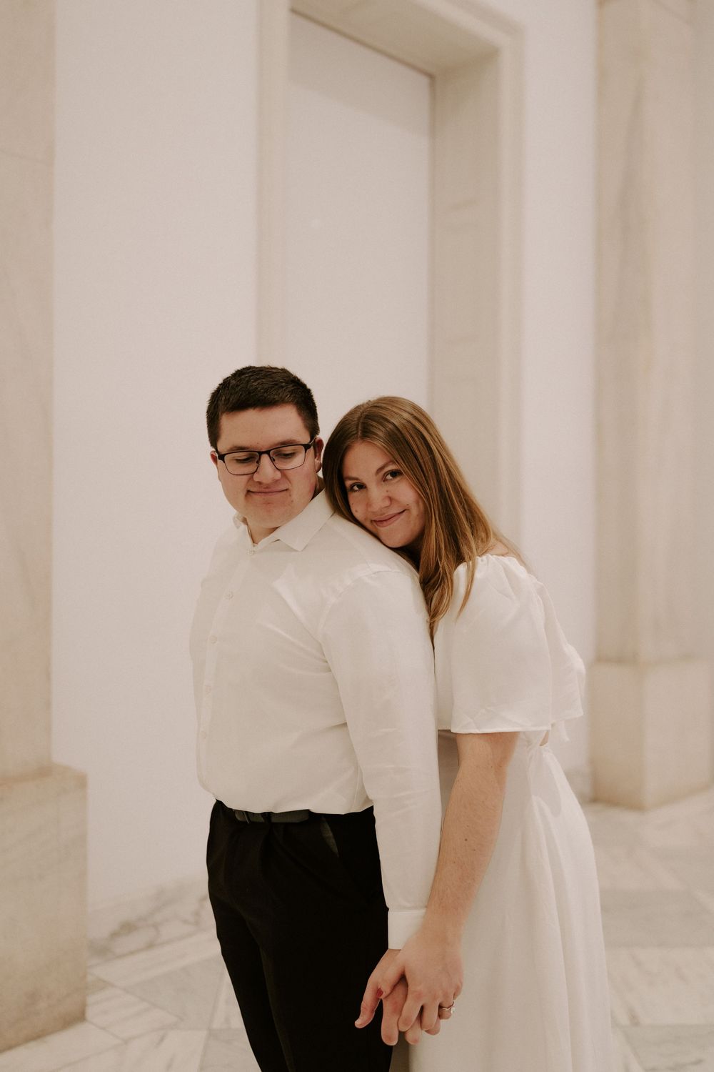 Engaged couple holding hands back-to-back in elegant marble building, woman in white dress smiling at camera.