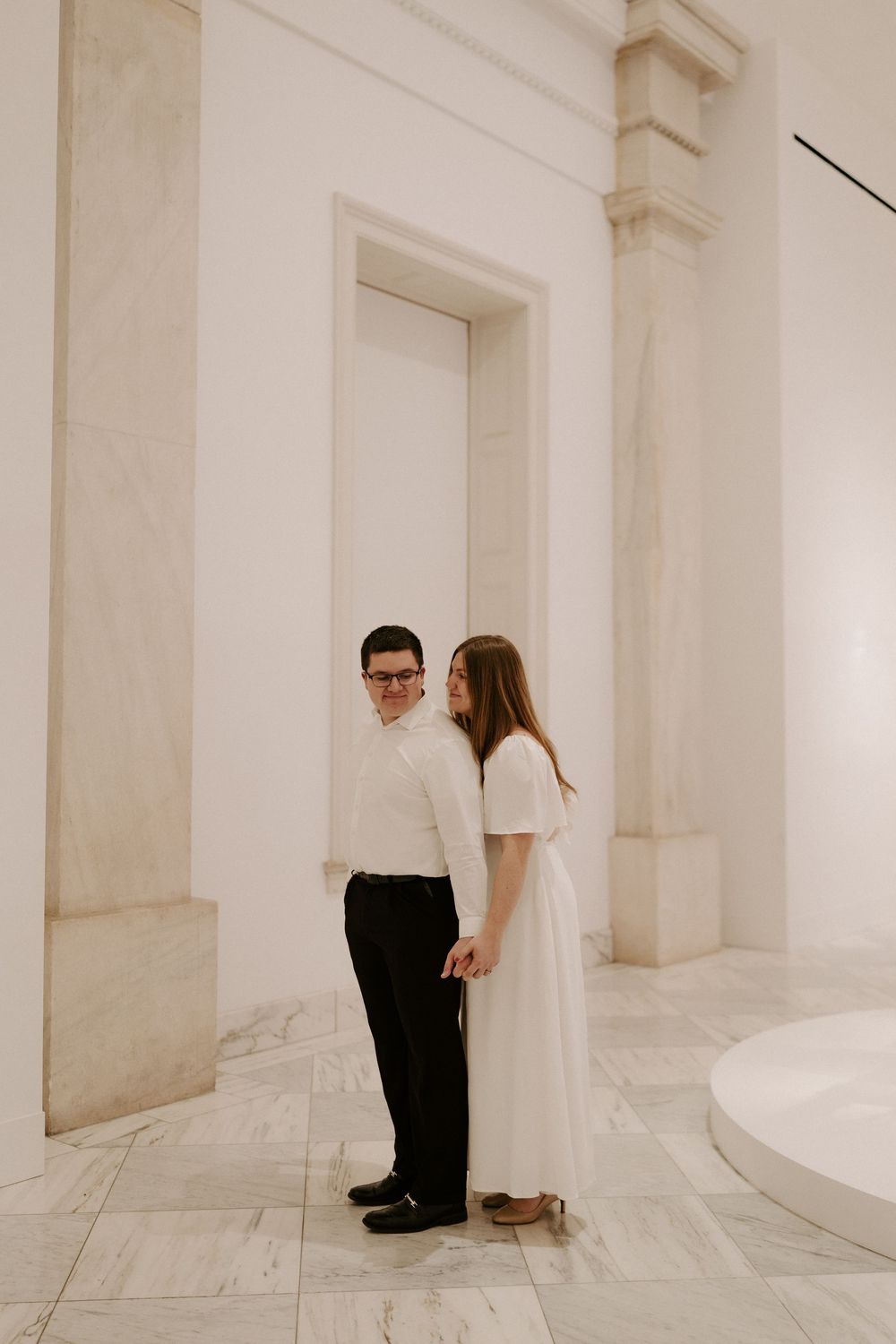 Couple holding hands and gazing at each other in a bright marble corridor with tall white columns.