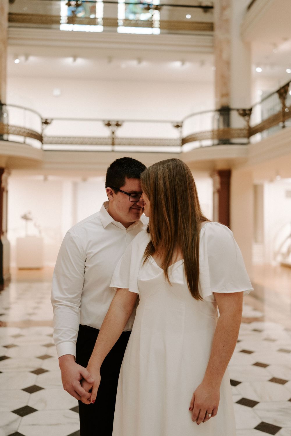 Romantic couple sharing an intimate moment in a grand marble hall with decorative balconies.