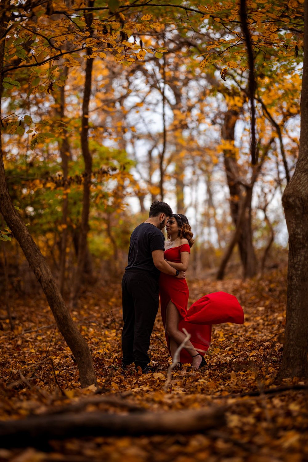 A couple dances together in a romantic autumn forest setting surrounded by fallen orange leaves and trees.