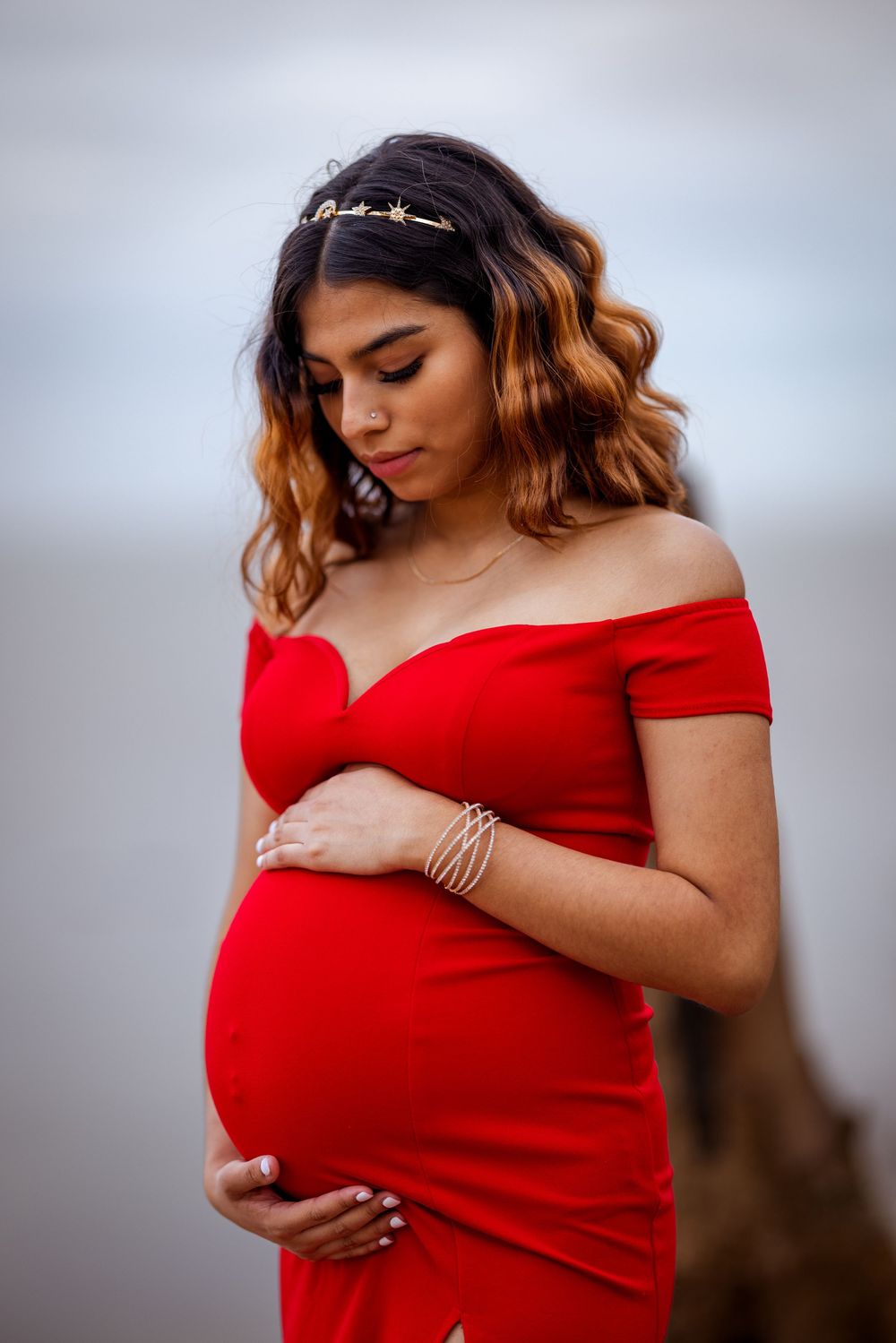 A person in a vibrant red off-shoulder dress poses gracefully against a soft gray background in a maternity photoshoot.