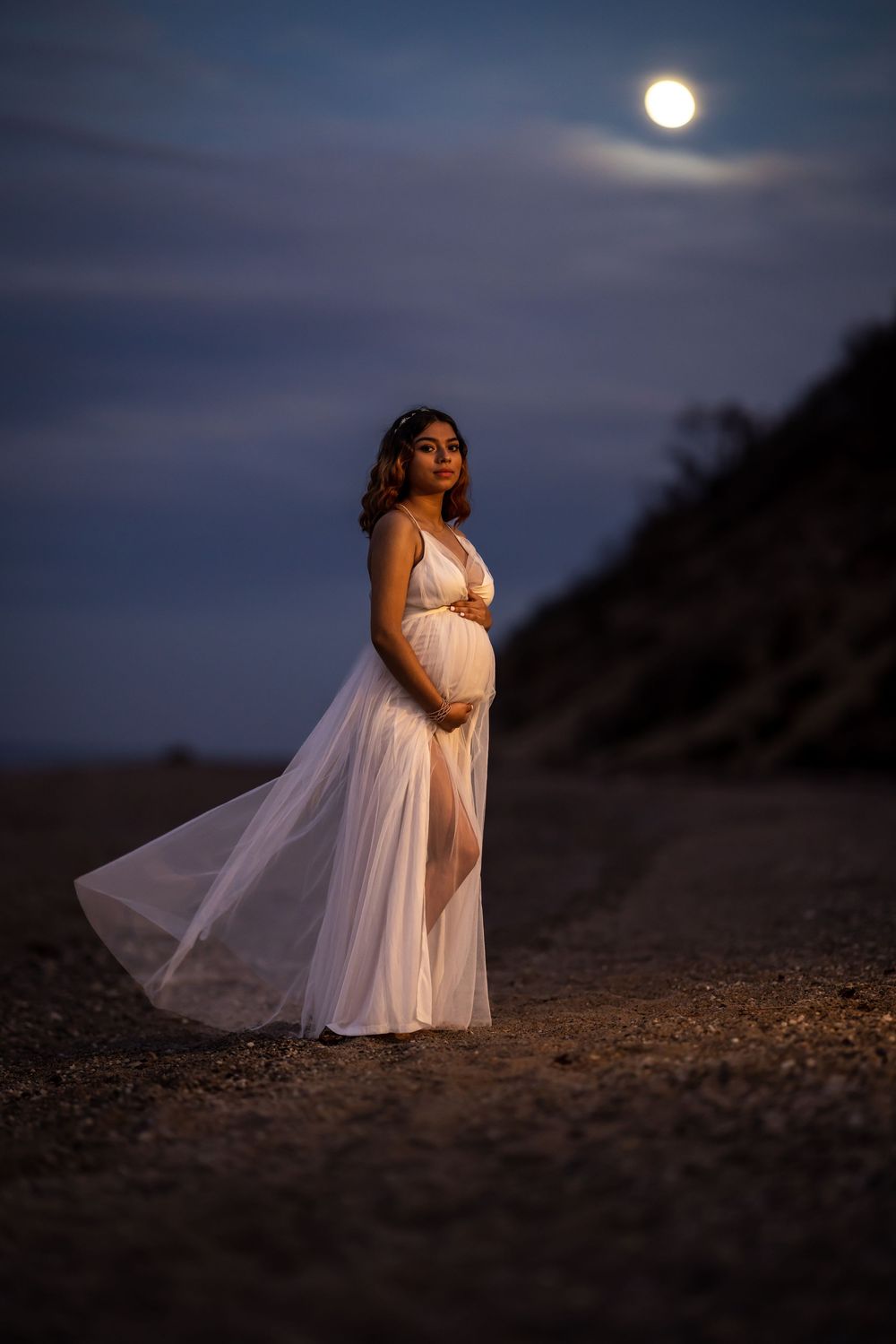 A figure in a flowing white dress poses on a beach at dusk with a full moon glowing in the dark blue sky.