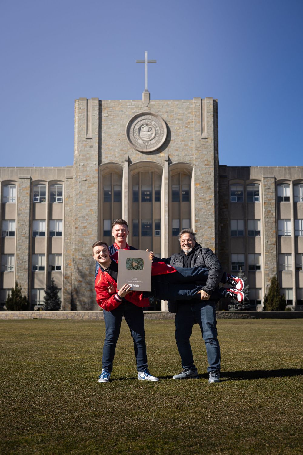 People posing playfully on lawn in front of large stone institutional building with cross on top.