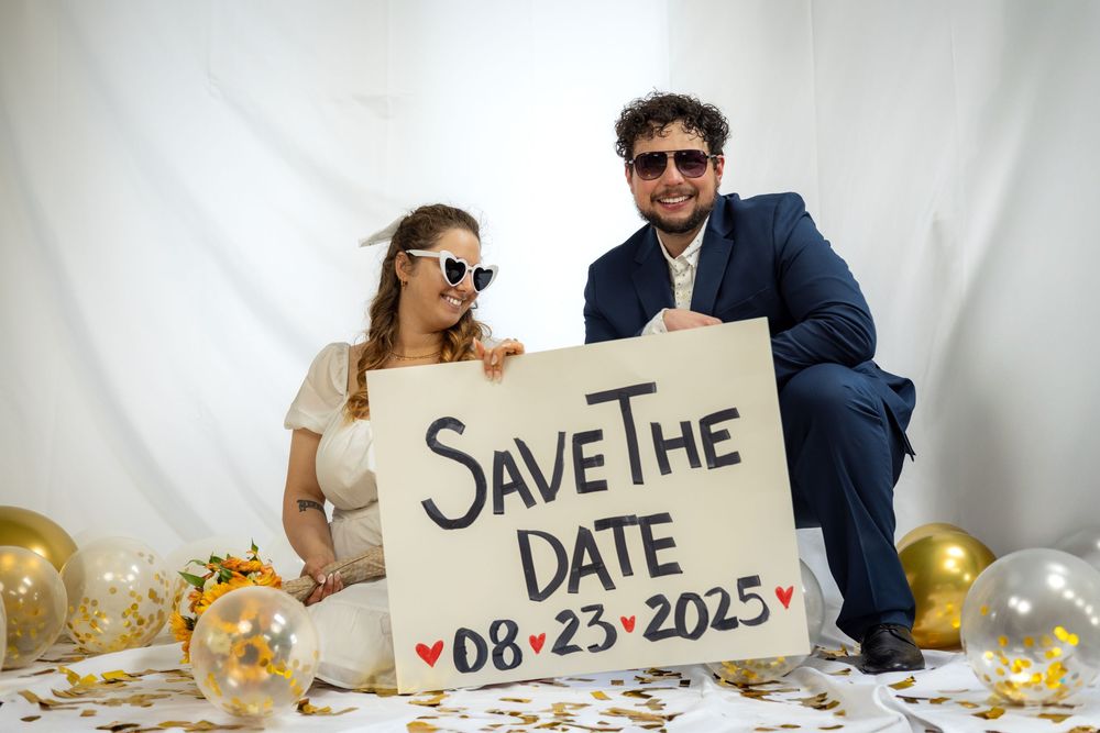 A couple holds up a series of signs spelling out a save the date message with gold decorations in the background.