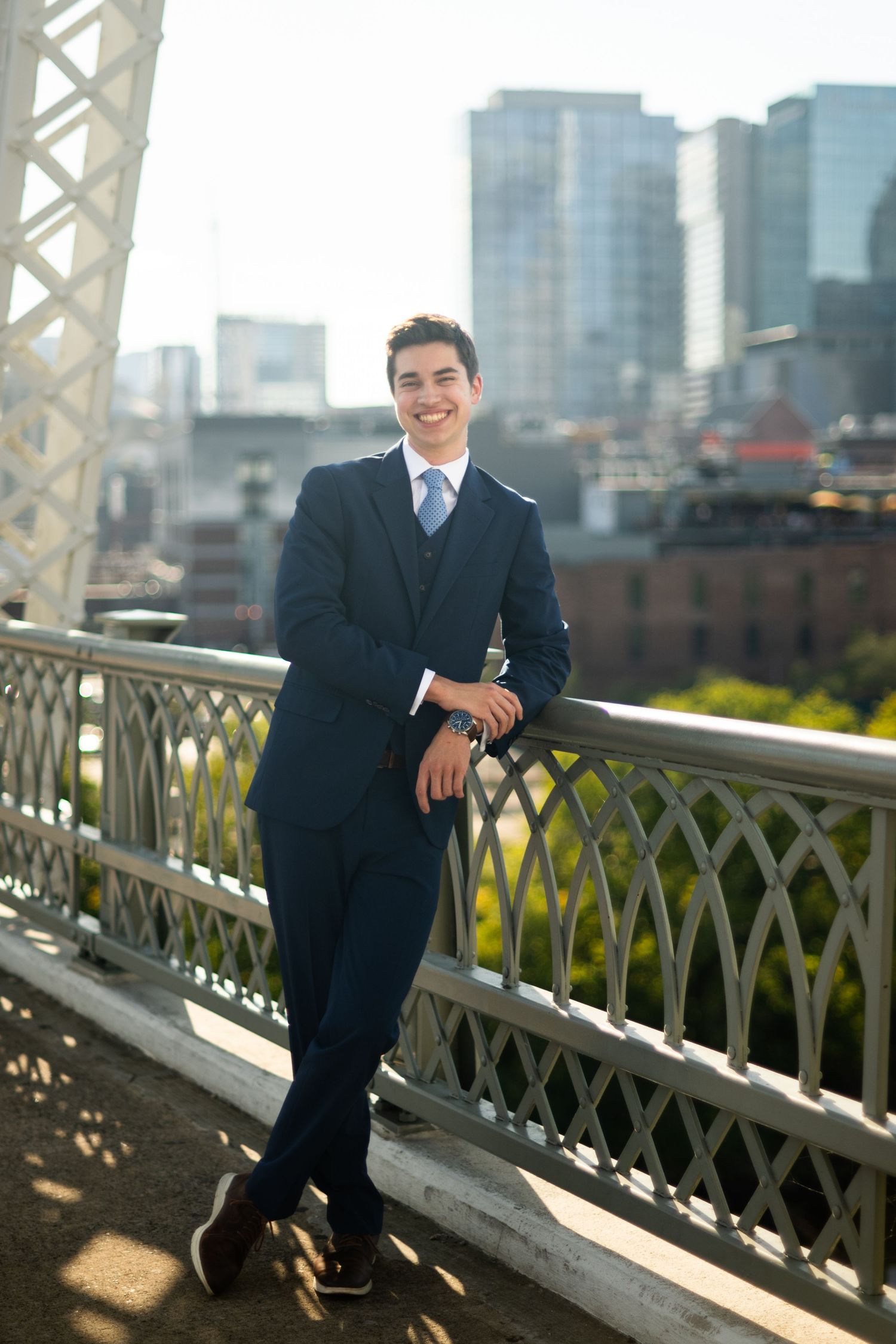 Professional portrait of a person in a dark suit posing on a decorative bridge railing with city buildings in background.