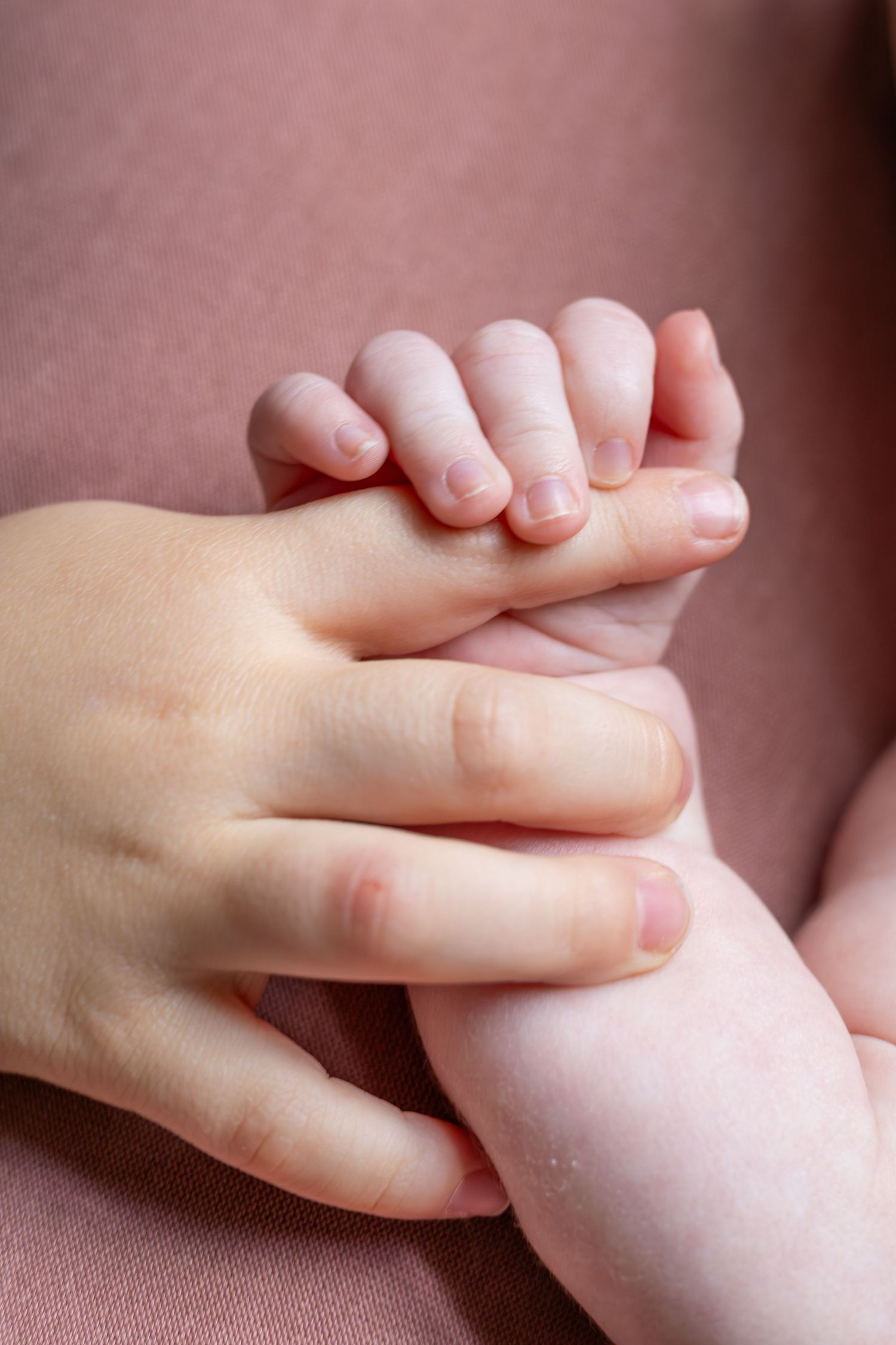 Close-up photo of a baby's tiny hand grasping an adult's finger against a soft pink background.