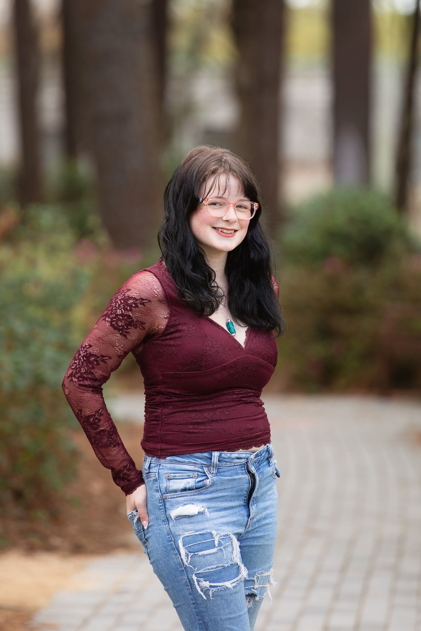 Young woman in burgundy lace top and ripped jeans smiling outdoors among trees on a brick pathway at WRAL Azalea Garden
