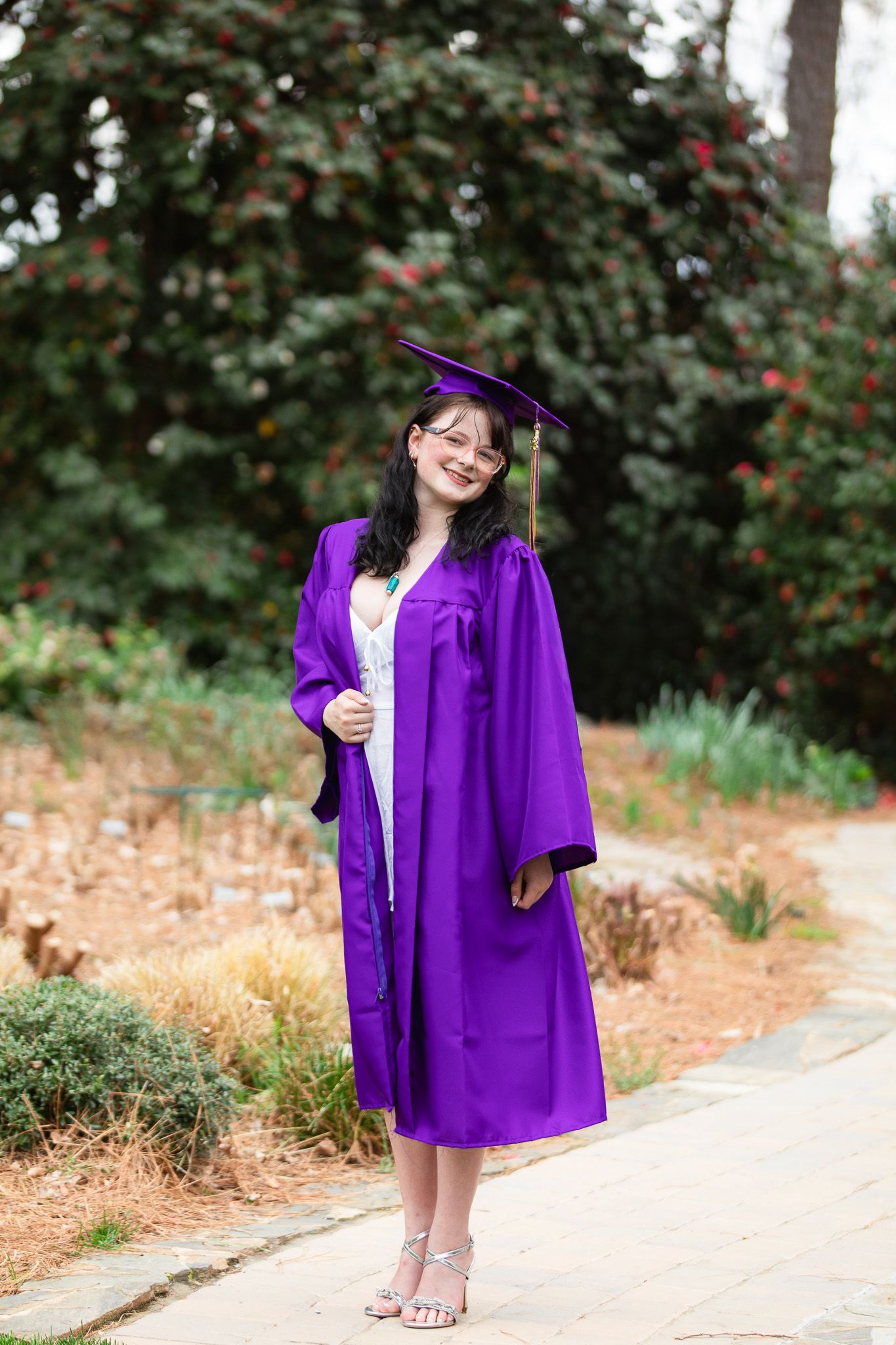 A young woman smiles in a purple graduation cap and gown, posing outdoors on a garden path at WRAL Azalea Garden