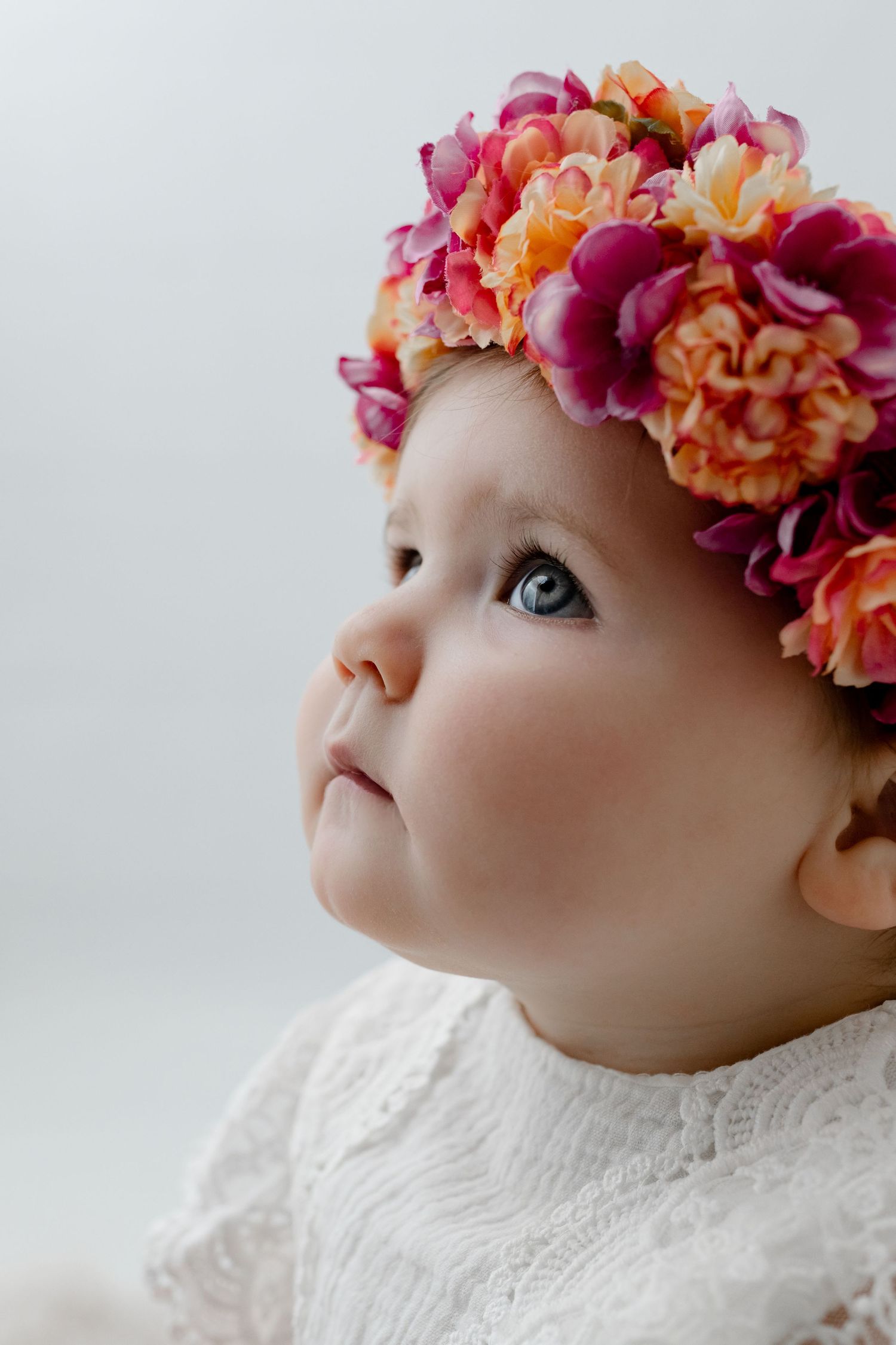 Delicate portrait of a young child wearing a vibrant pink and orange floral crown and white lace dress, gazing upward.
