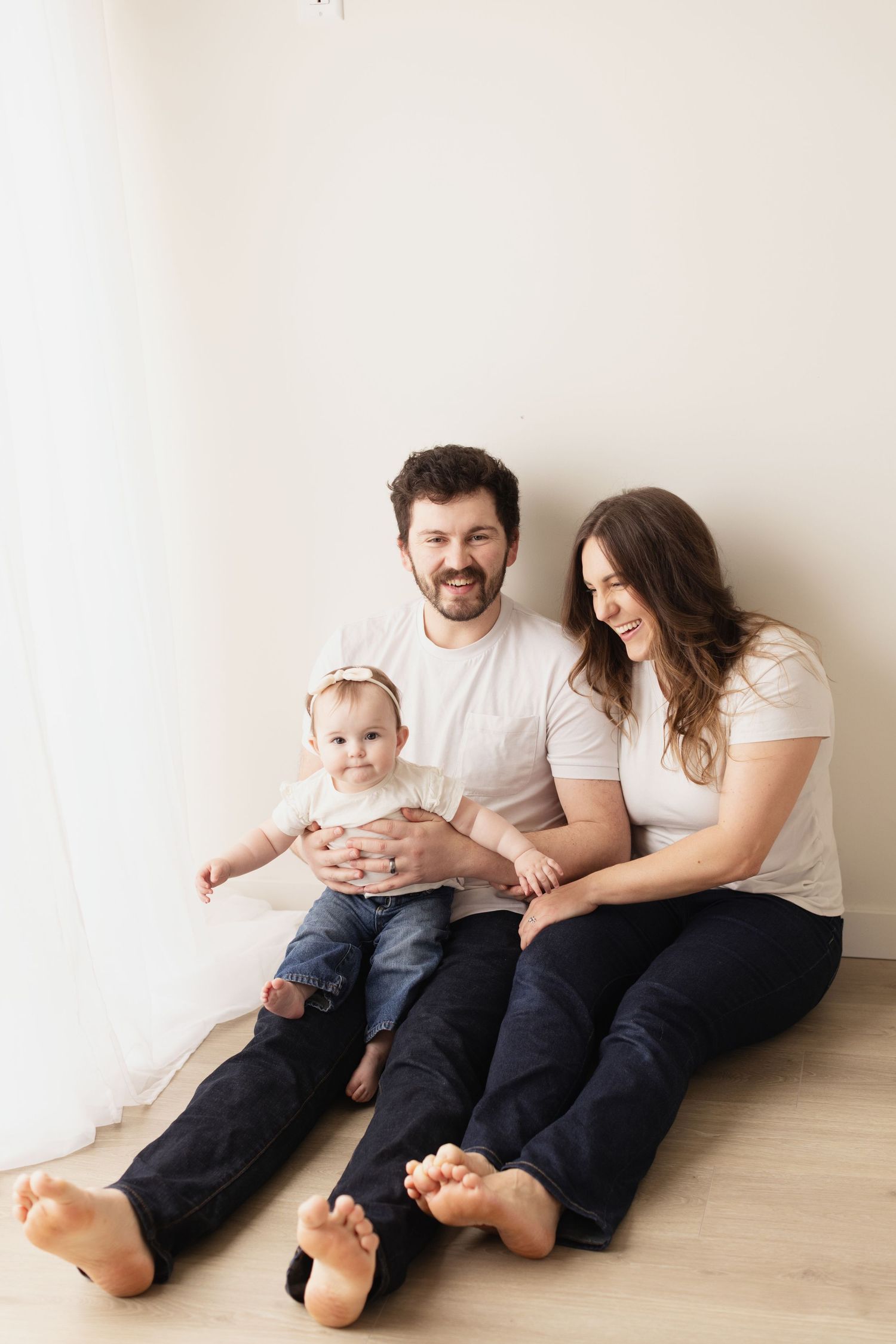 A family of three sits together on a wooden floor wearing white shirts and dark pants while sharing joyful moments. Elizabeth Lang Photograp