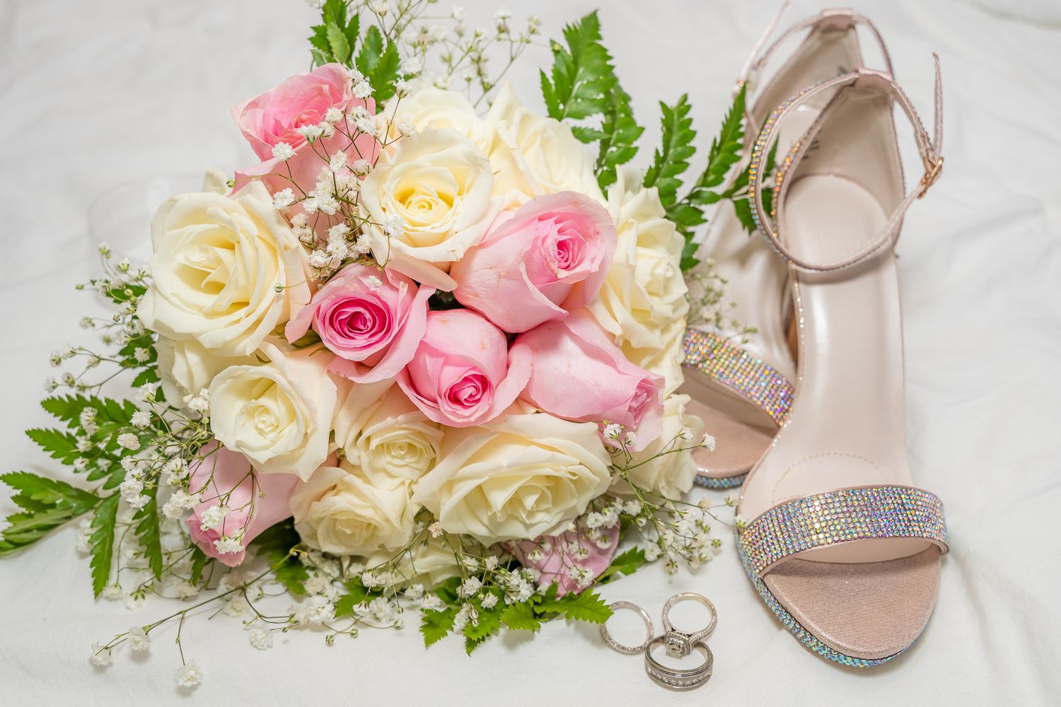 Delicate pink and white rose wedding bouquet displayed with silver rings and beige sandals on white linen.