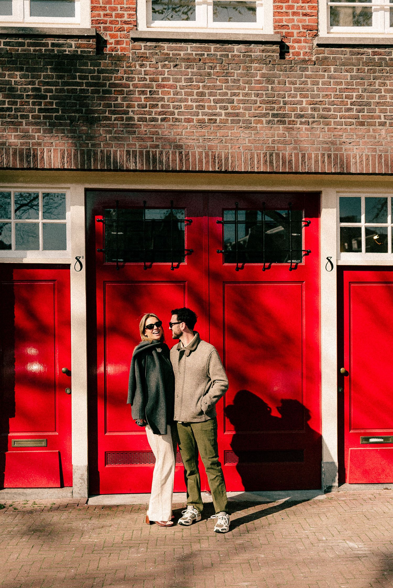 Couple standing together in front of vibrant red doors on a brick building, enjoying a sunny day outdoors.