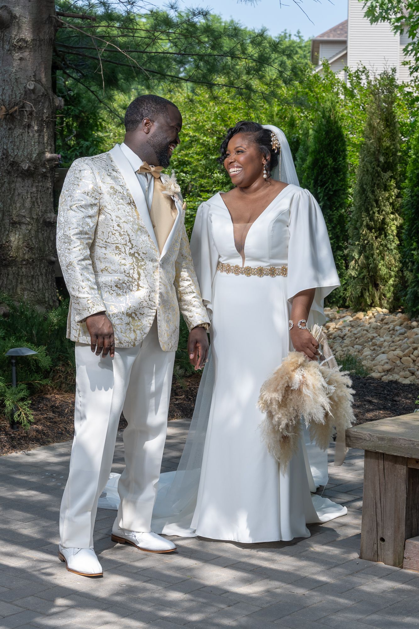 A joyful Black couple in elegant wedding attire, groom in gold brocade suit, bride in white gown with veil, outdoors.